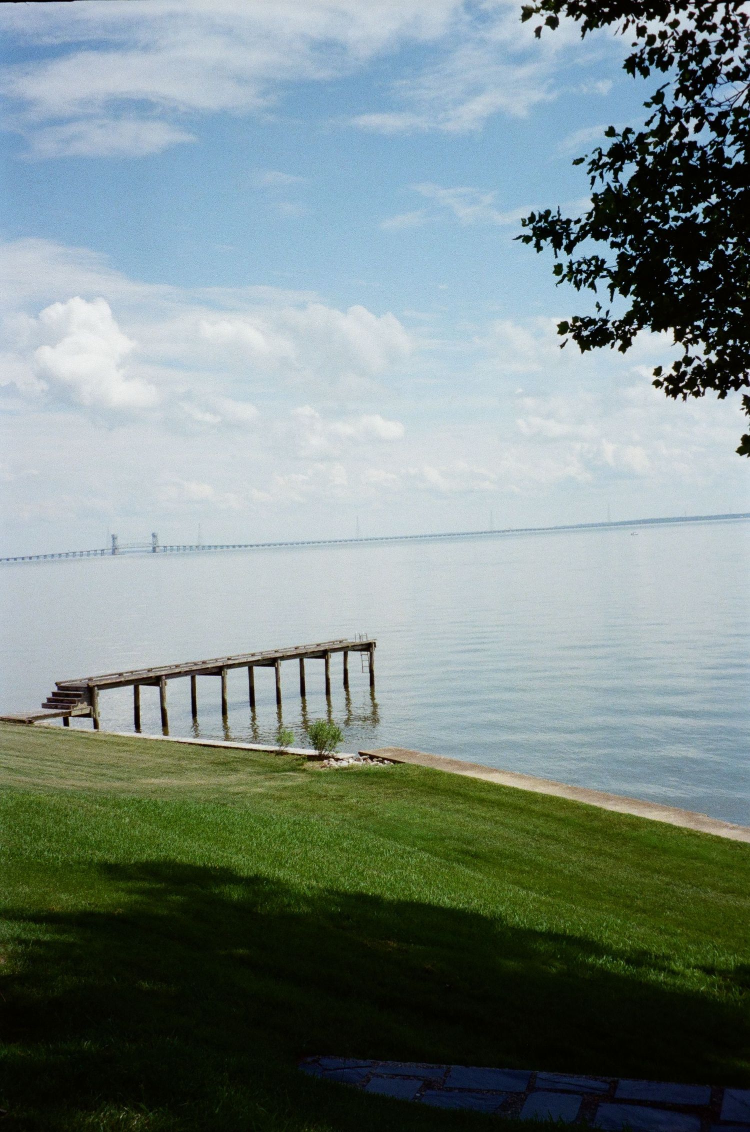 A wooden pier extends into the calm waters of the Chesapeake Bay with the Bay Bridge visible in the distance.