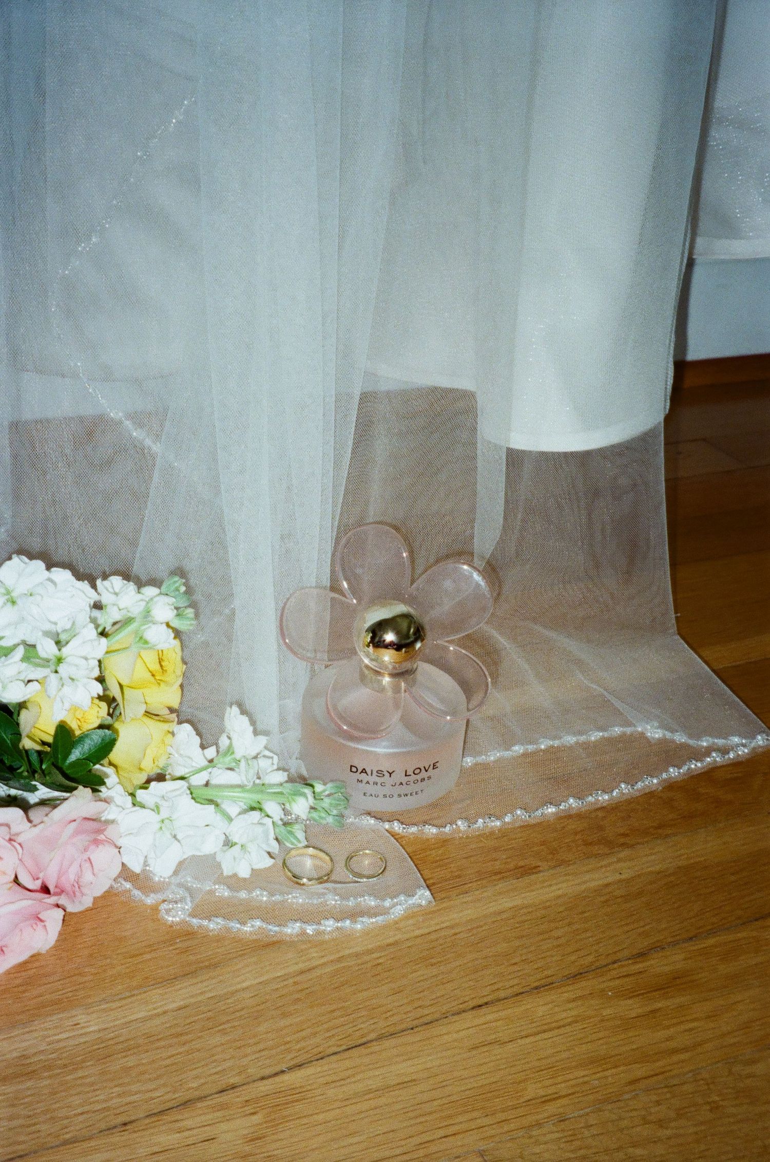 Pink perfume bottle with flower-shaped cap sits beside white and yellow flowers on a wooden floor with sheer curtains.