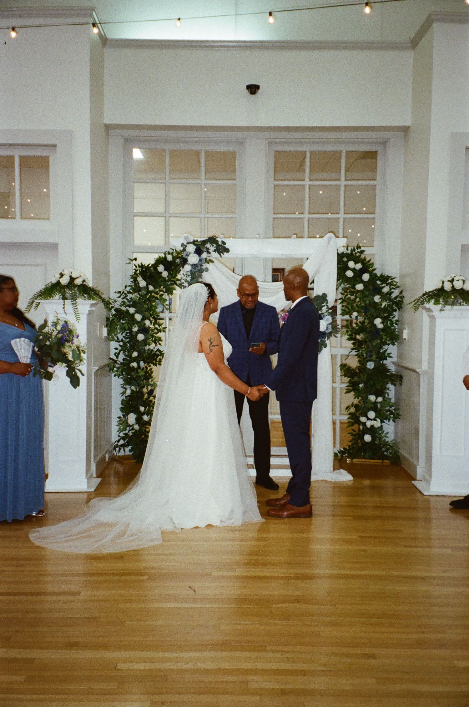 A wedding ceremony takes place under a beautiful floral arch with white flowers against French doors in an indoor venue.