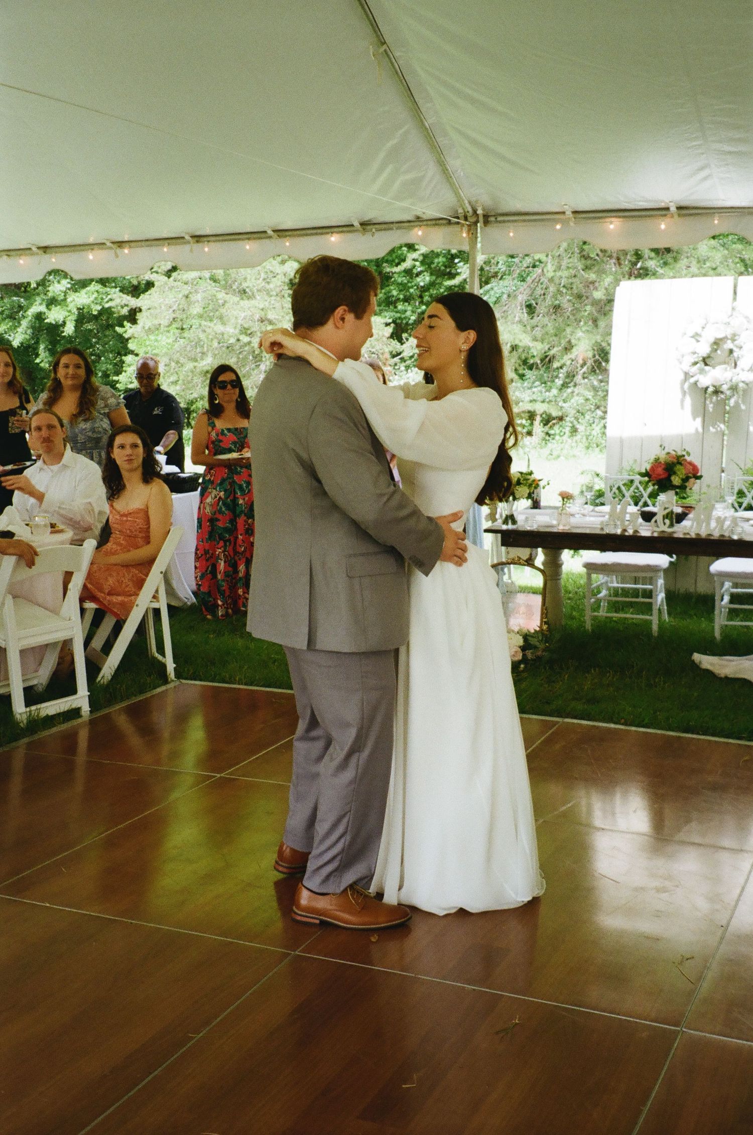 A couple shares their first dance under an elegant white tent during a backyard wedding reception.