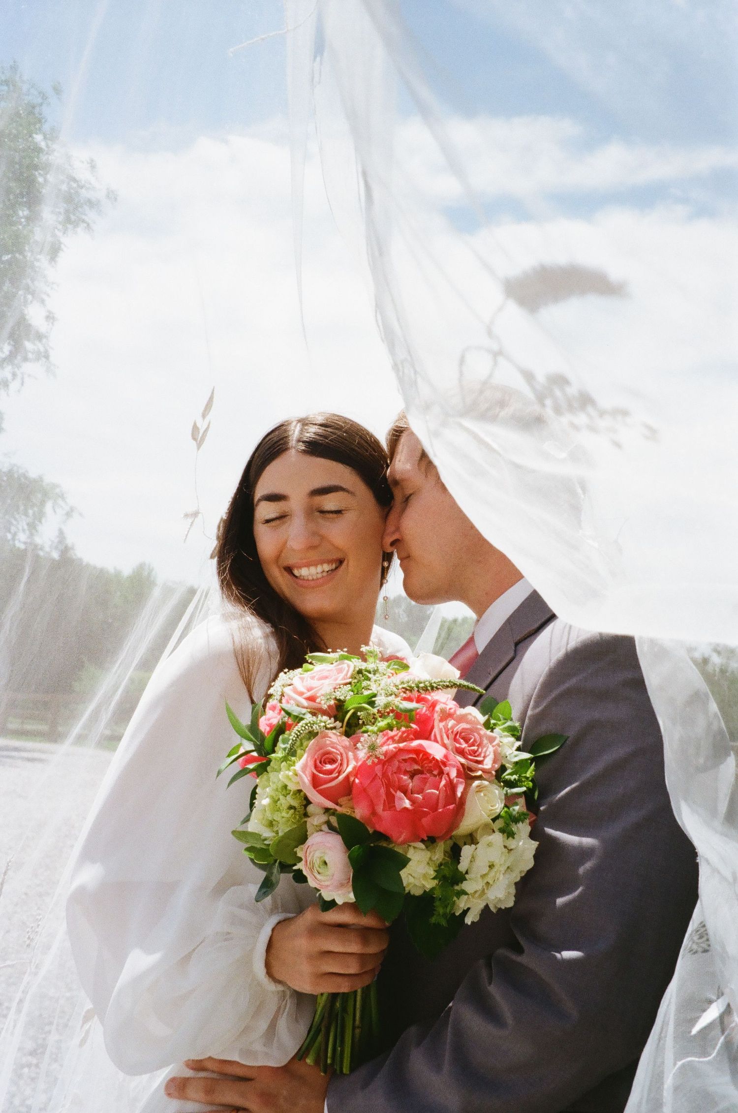 A romantic wedding moment under a flowing white veil with a coral rose and white flower bouquet.