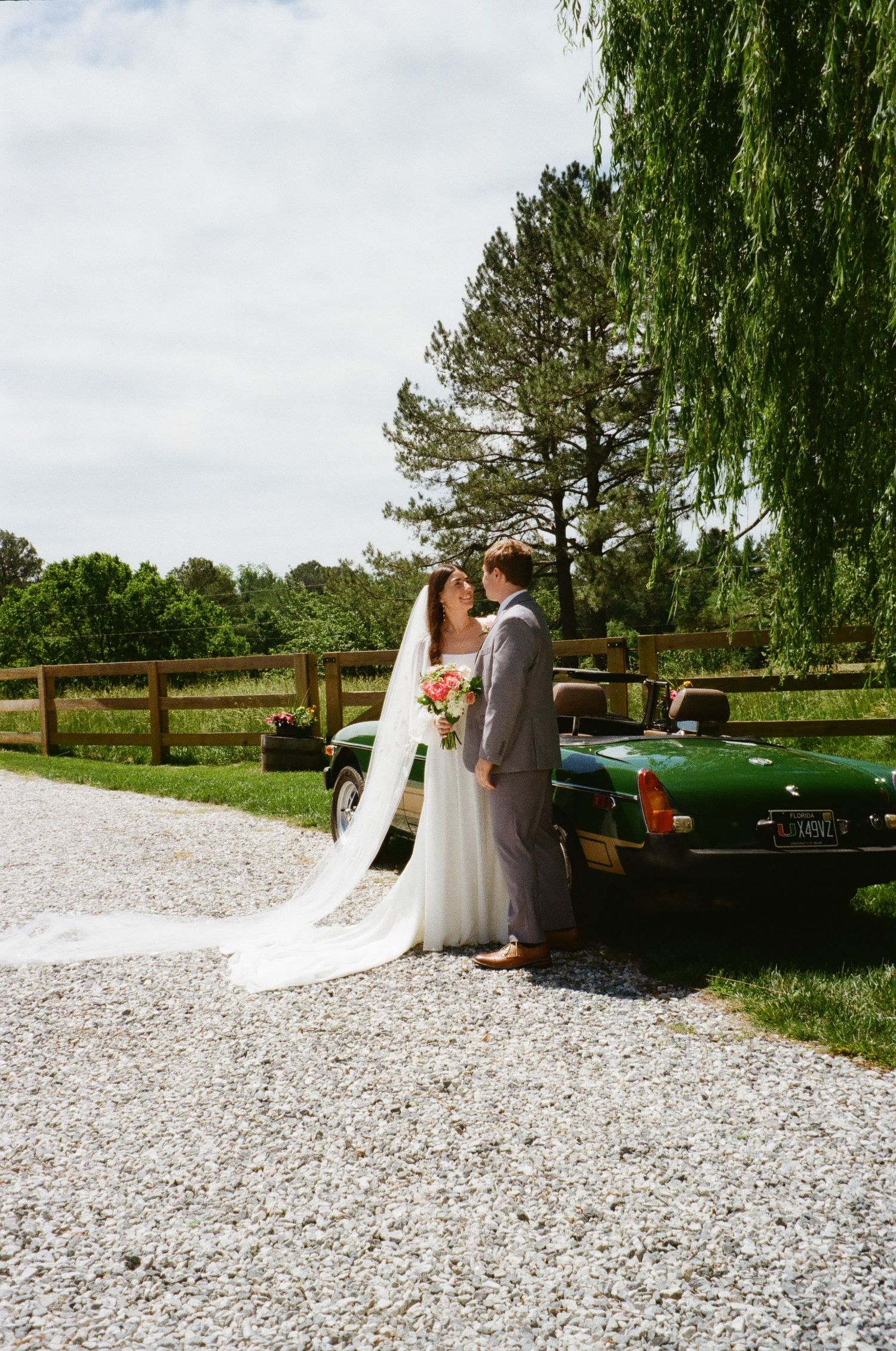 Bride and groom share a romantic moment beside a vintage green car on a gravel path, surrounded by lush greenery.