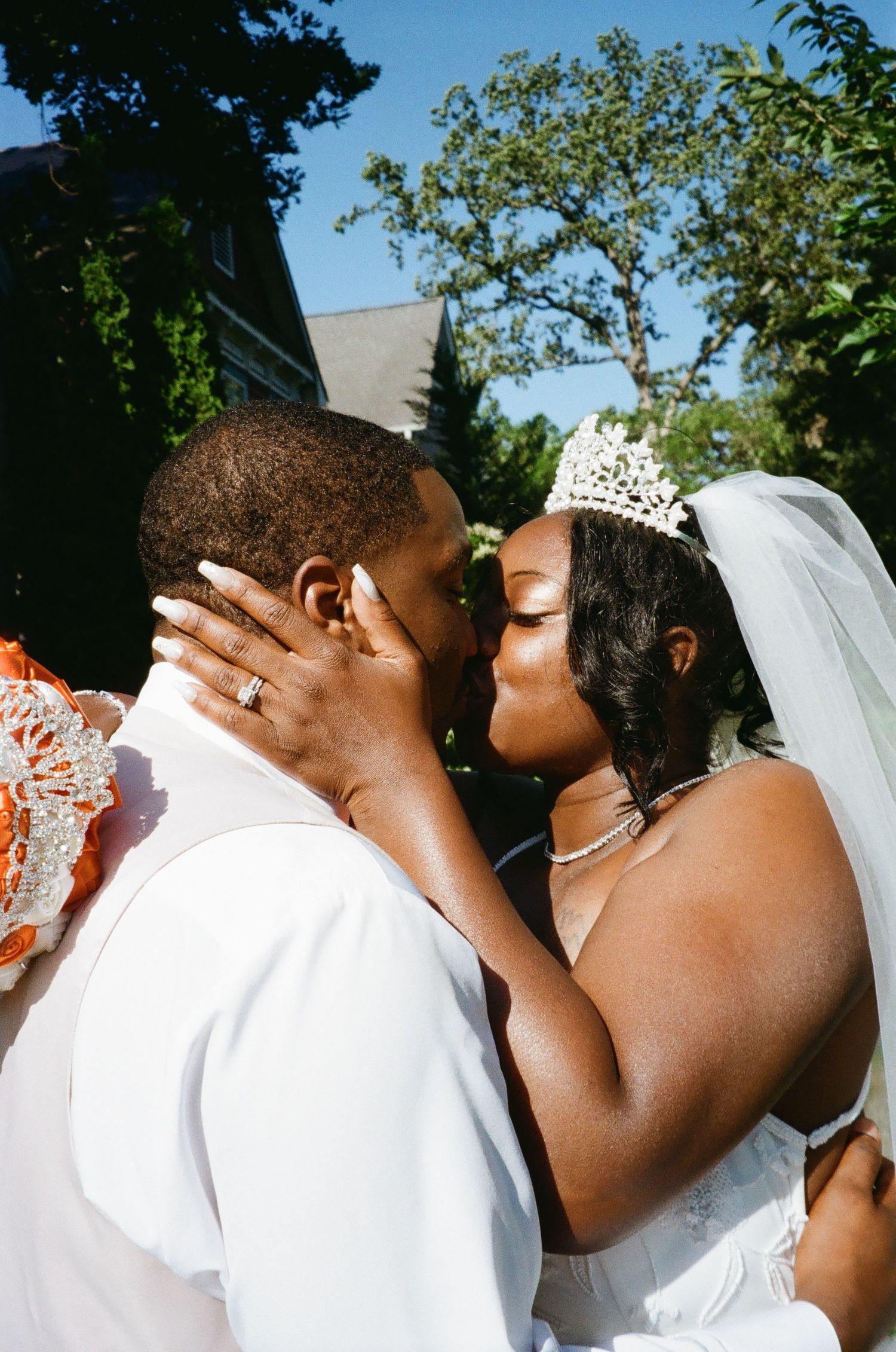A tender wedding day kiss captured outdoors with the bride wearing a tiara and veil and groom in white attire.