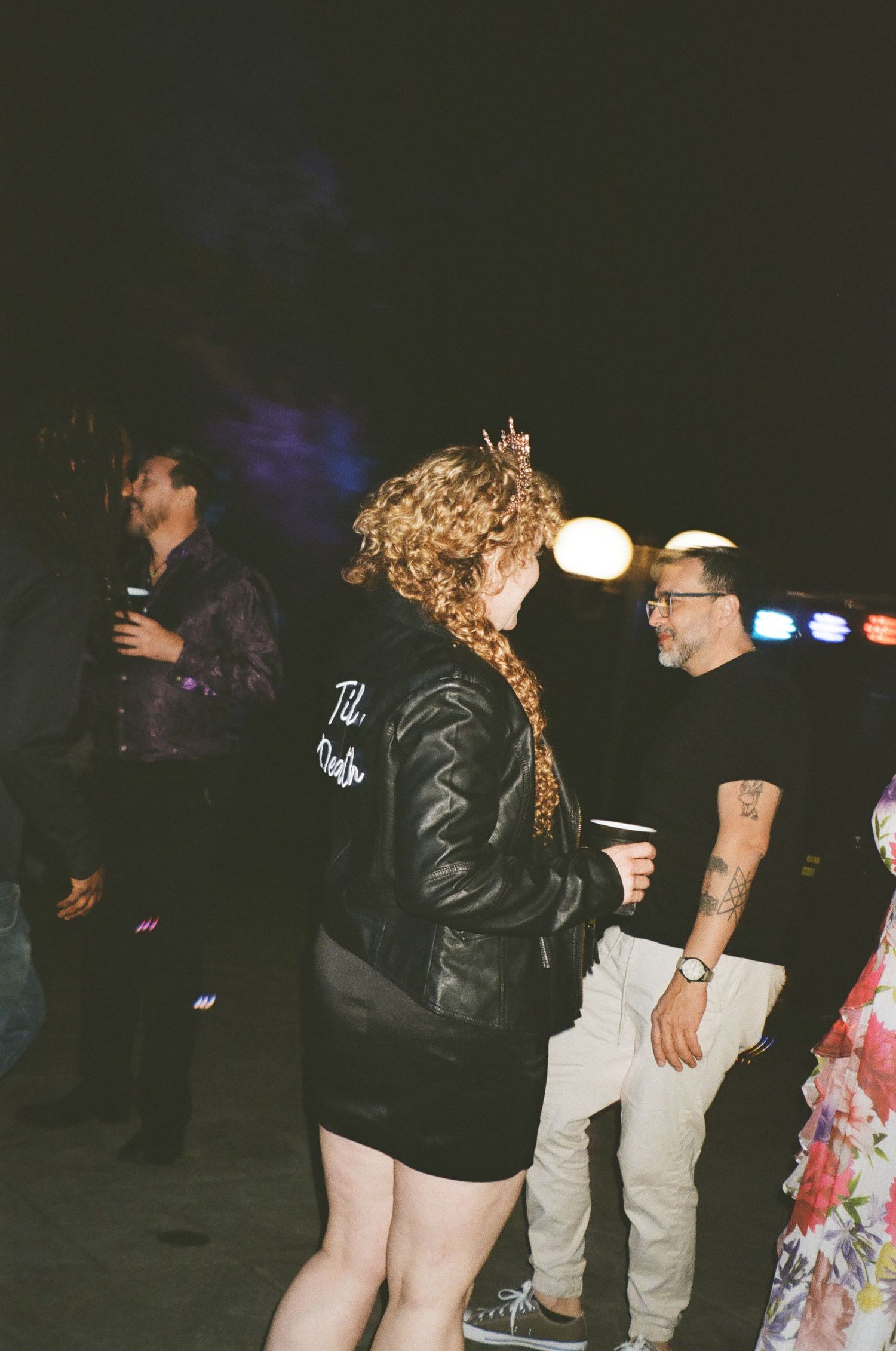Group of people hanging out at night with curly-haired person in black jacket in focus at outdoor gathering.