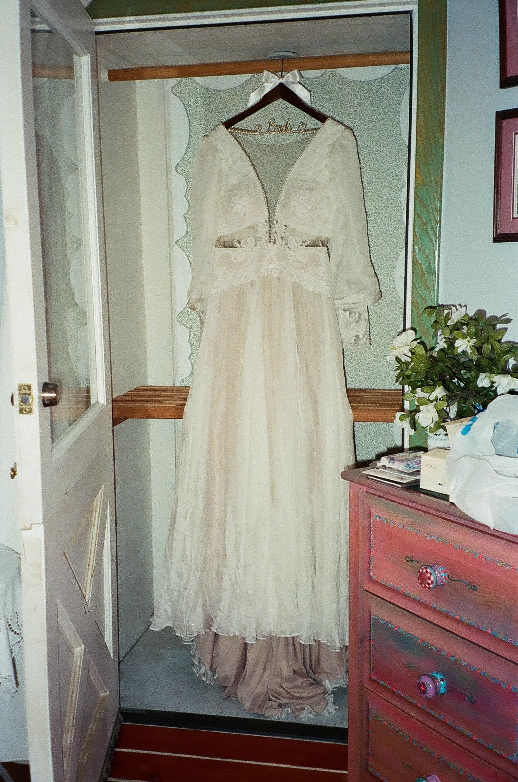 A vintage white lace wedding dress with long sleeves hangs in a wooden wardrobe next to a red dresser.