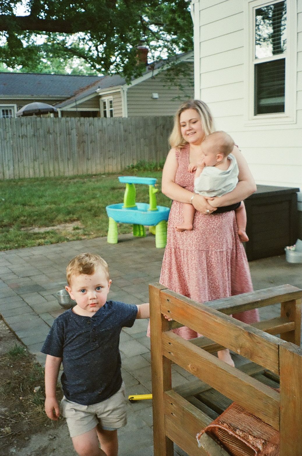 Family outdoors on backyard patio with wooden bench and children's play table visible in the background.