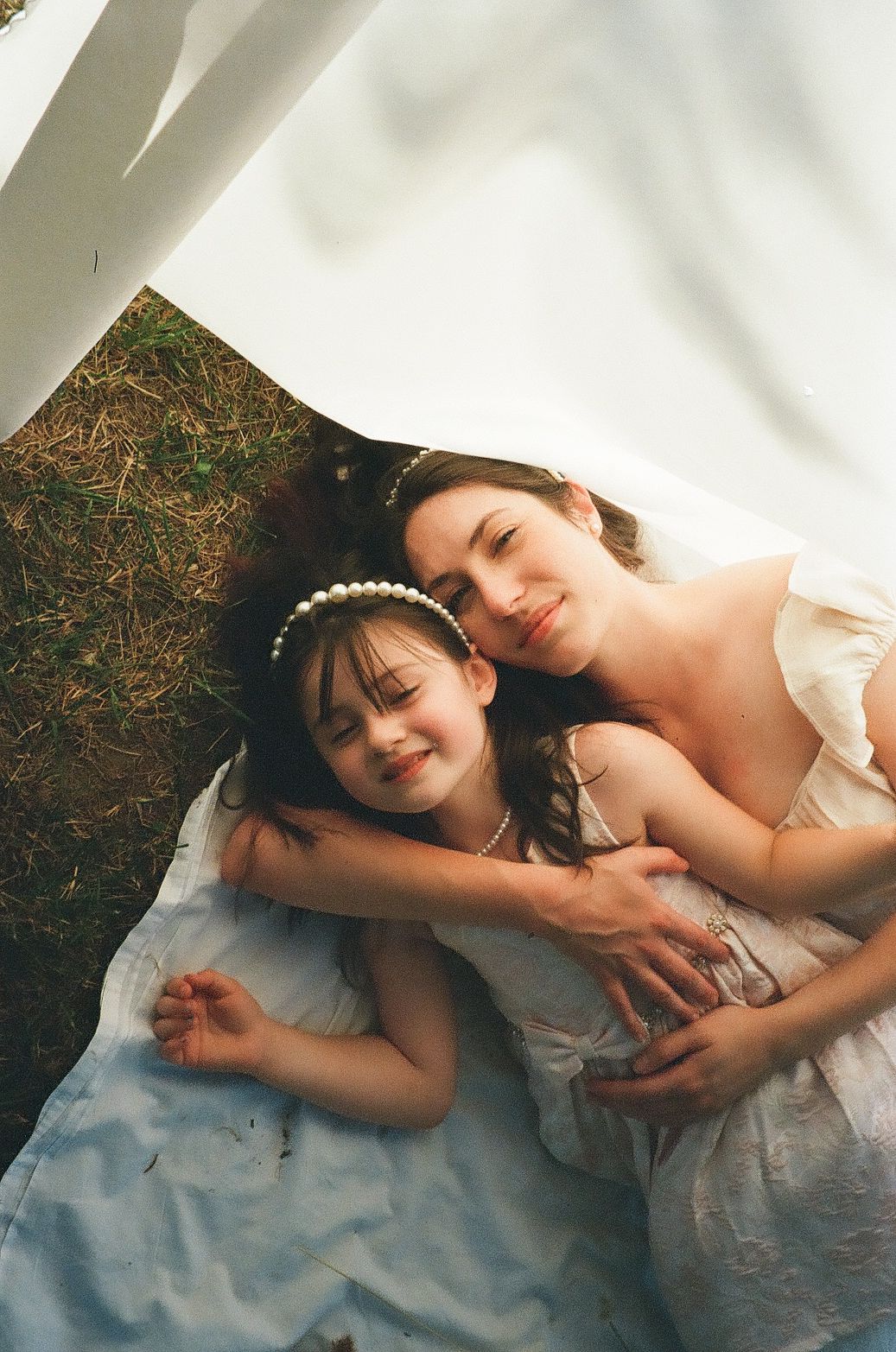 Two people in white dresses laying down on a blanket outdoors on a sunny day.