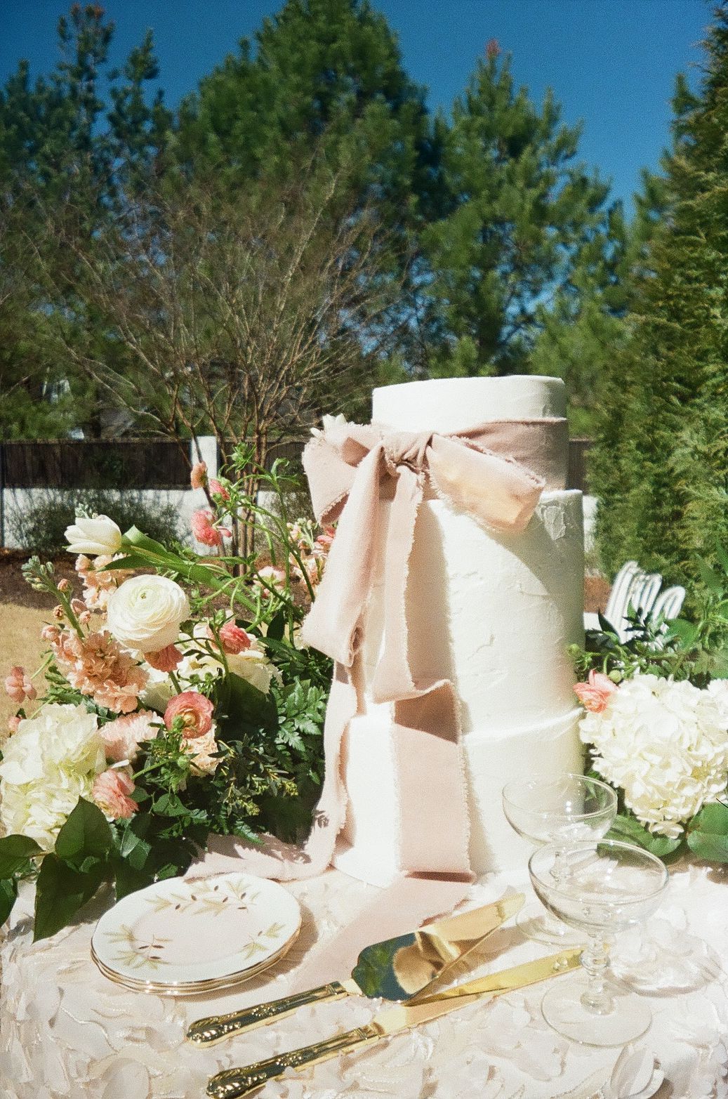 Elegant wedding table setting with white cake adorned with blush ribbon, surrounded by pink and white floral arrangements outdoors.