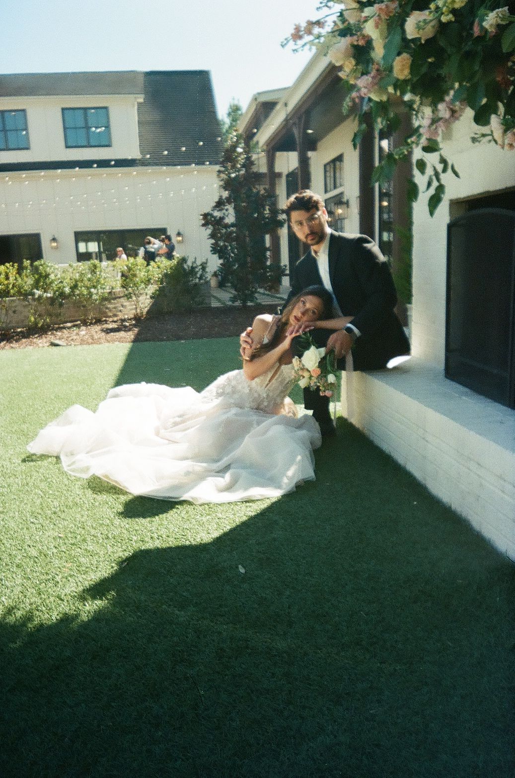 A couple in wedding attire shares a joyful moment on artificial turf outside a white building with string lights.