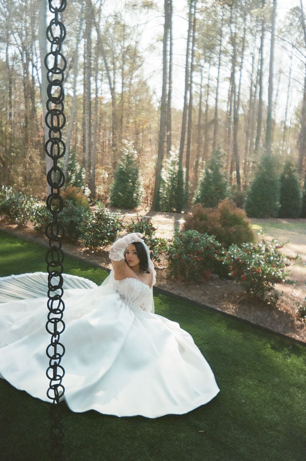 A bride in a flowing white gown twirls elegantly on a swing in a serene garden setting with tall pine trees.