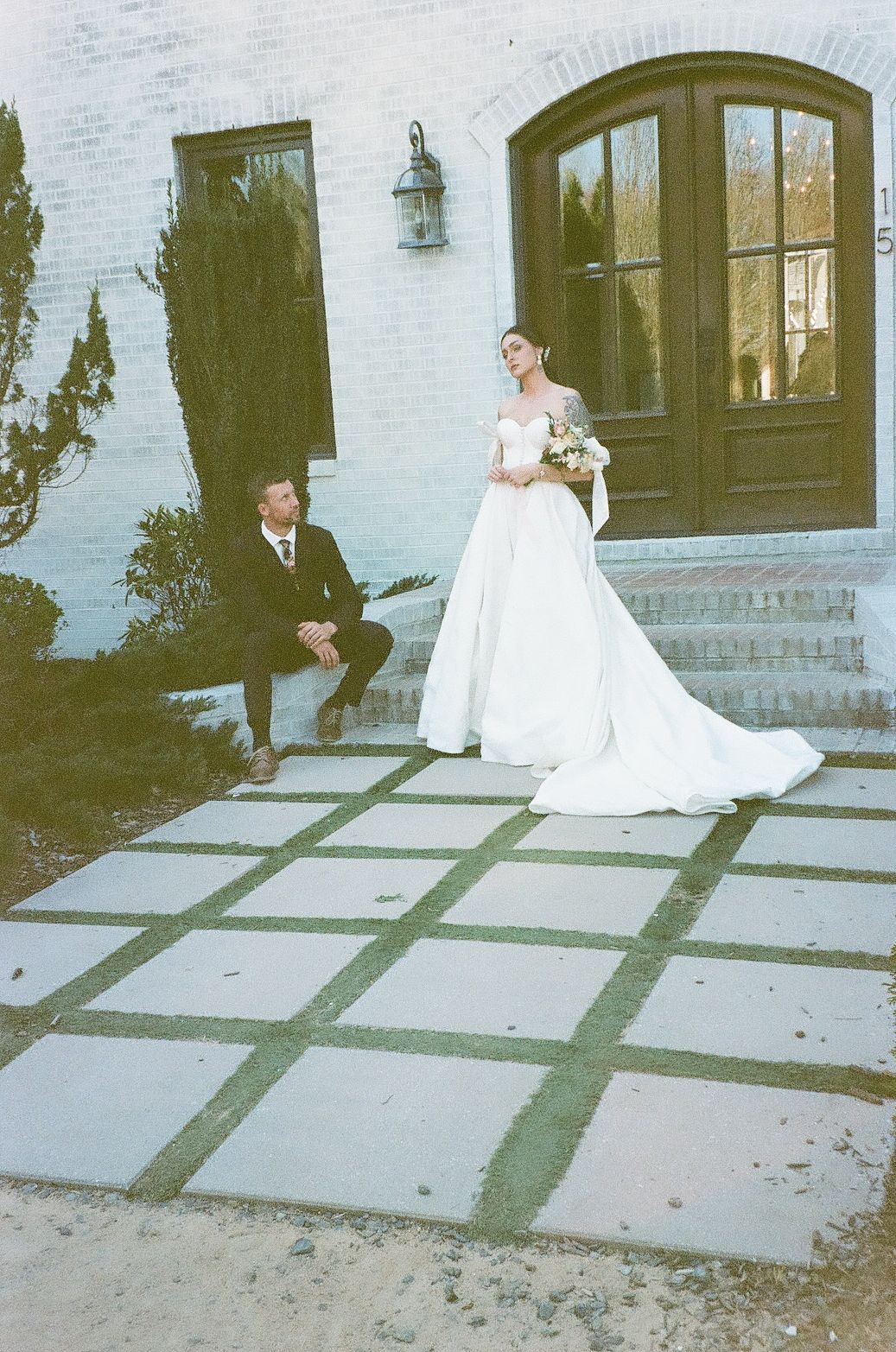 A wedding photo showing a couple on a tiled patio outside a white building with arched doorways and cypress trees.