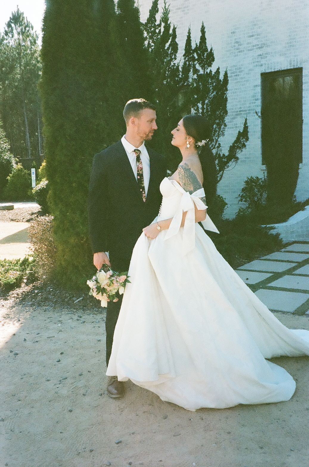 Couple in elegant wedding attire share an intimate moment in front of cypress trees outside a white building.