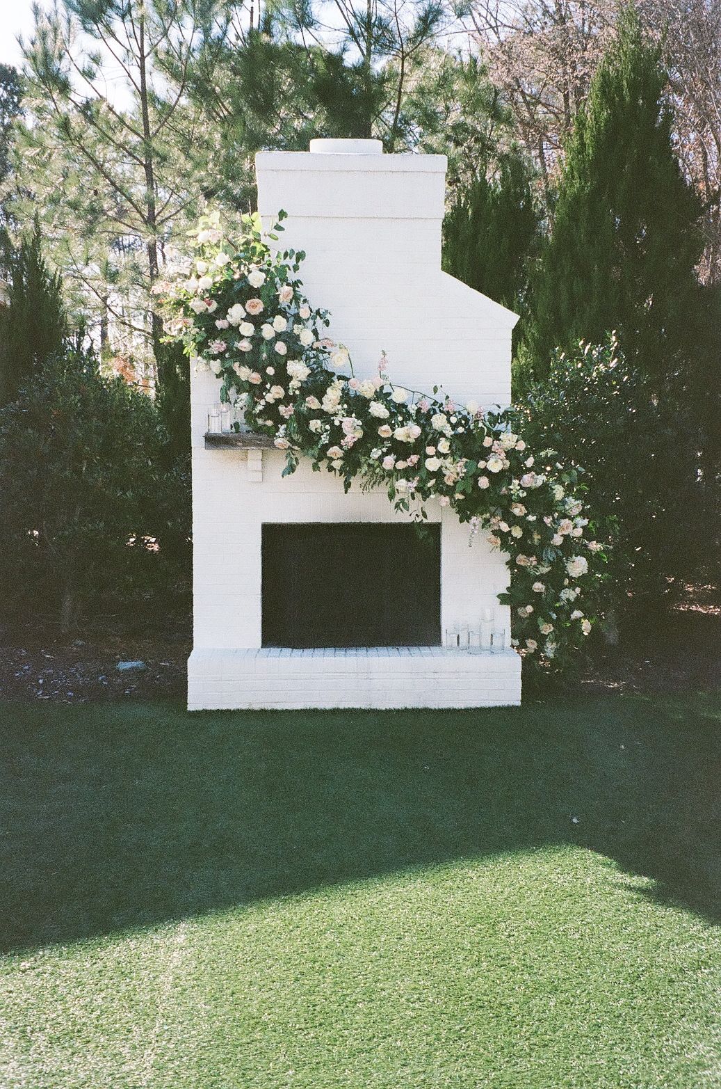 White outdoor fireplace decorated with cascading floral arrangements against evergreen trees.