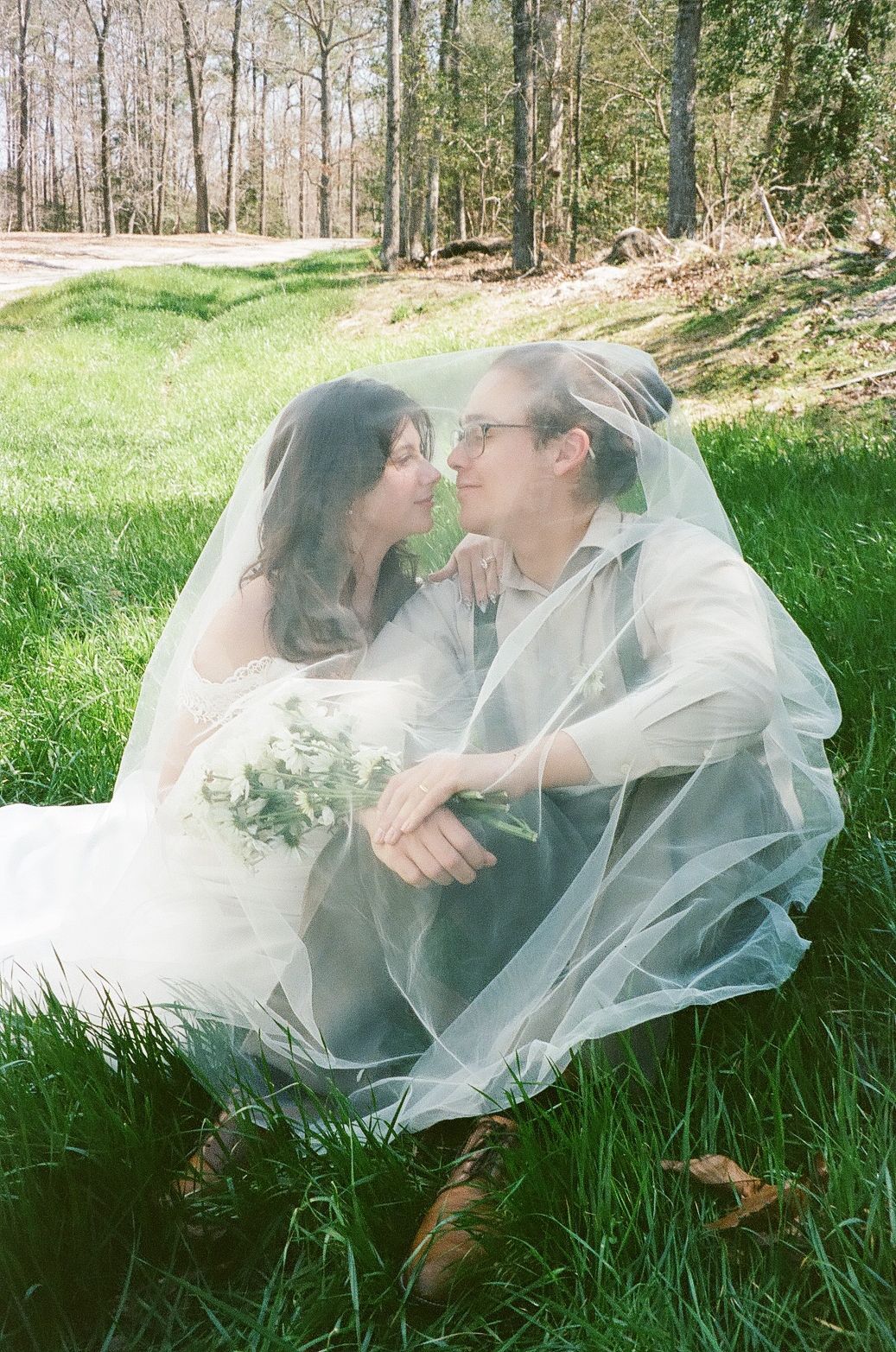 A couple wrapped in a wedding veil share an intimate moment while sitting on grass in a wooded setting.