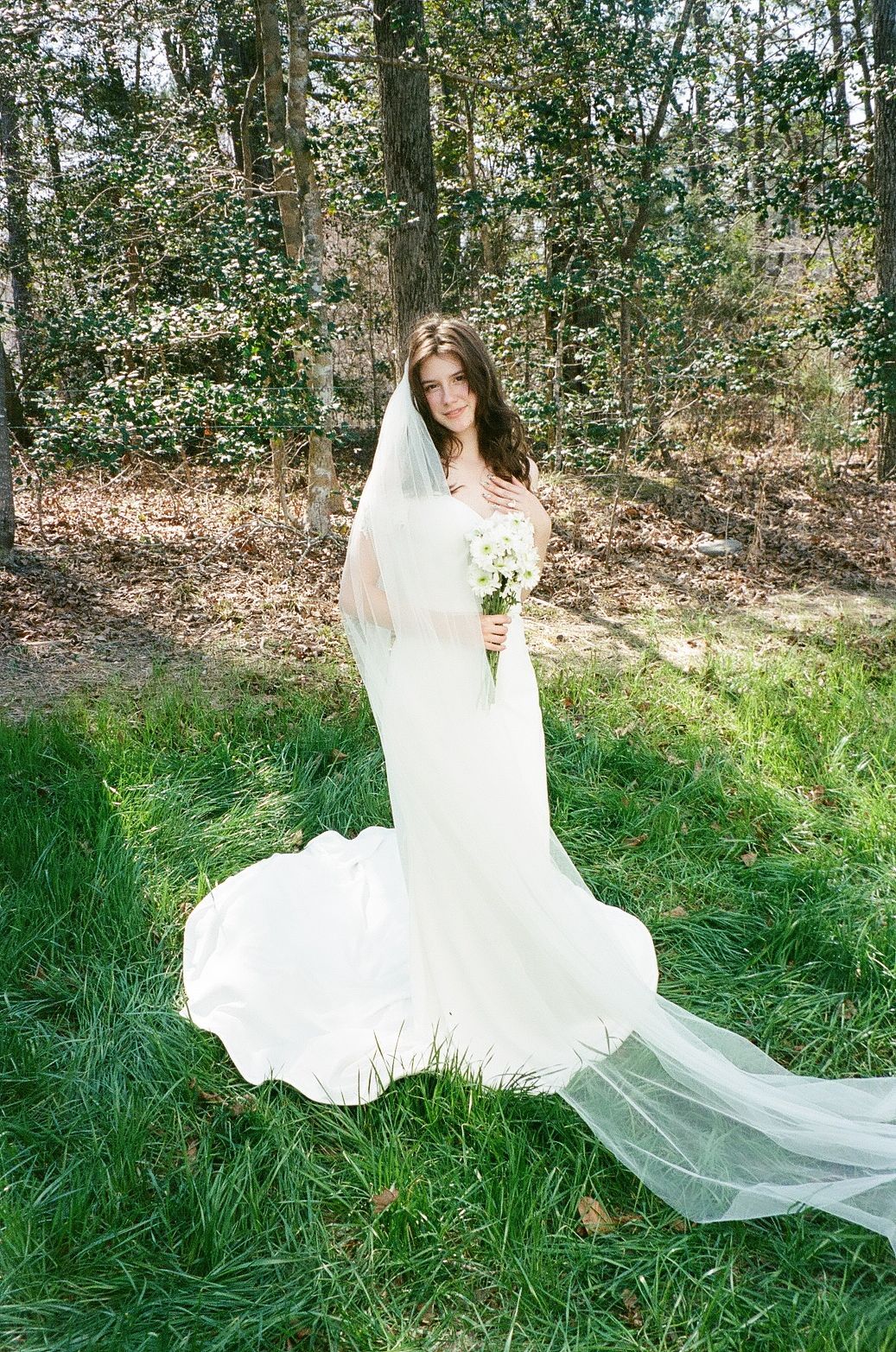 A bride in an elegant white lace gown with a flowing veil holds a bouquet while standing in a wooded outdoor setting.