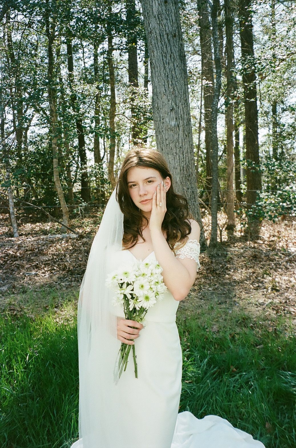 A bride in a white wedding dress holds a bouquet of white flowers while standing in a sunlit wooded area.
