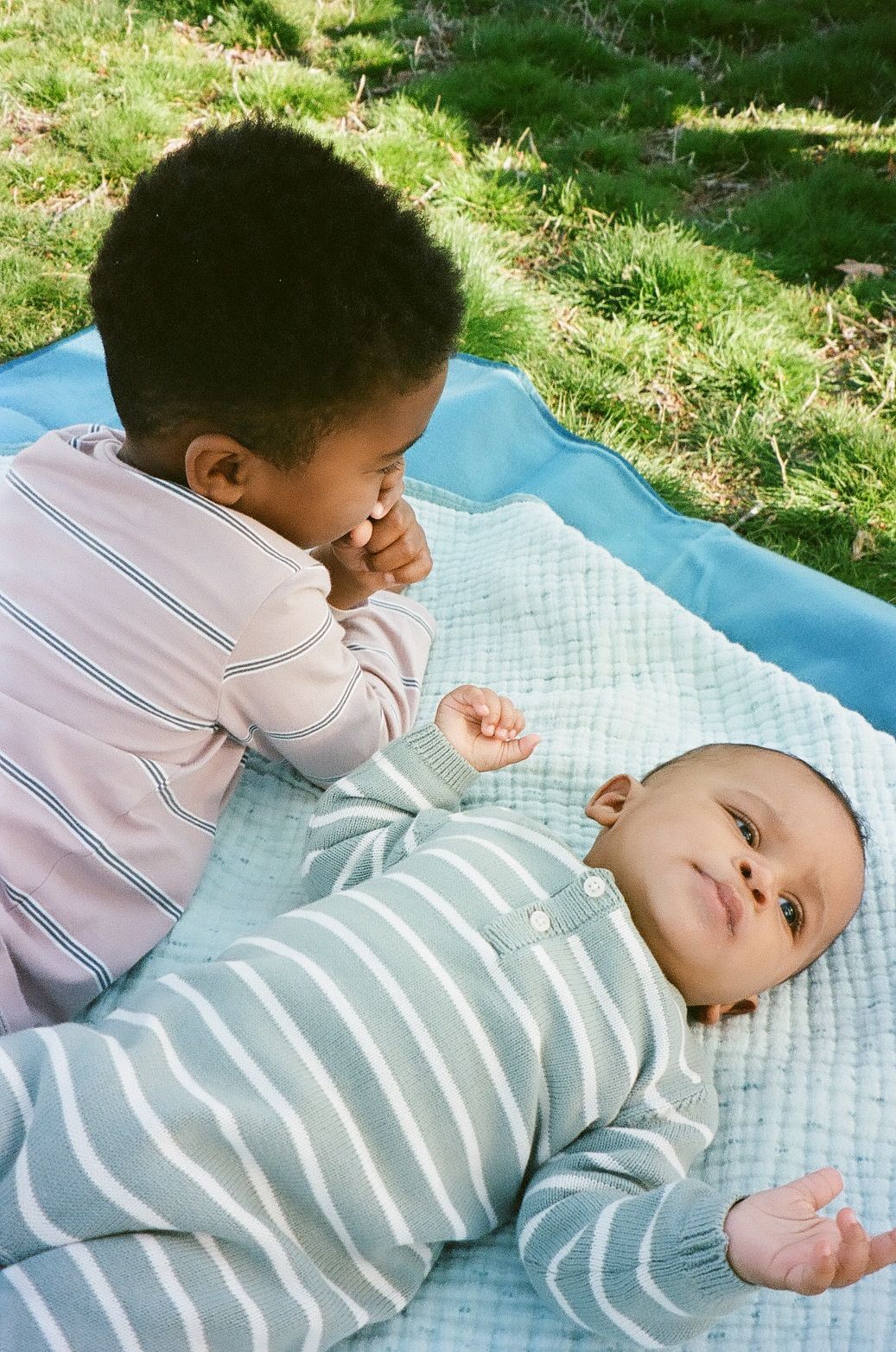 Two young children resting on a blue blanket outdoors, with one looking down at the other on a sunny day.