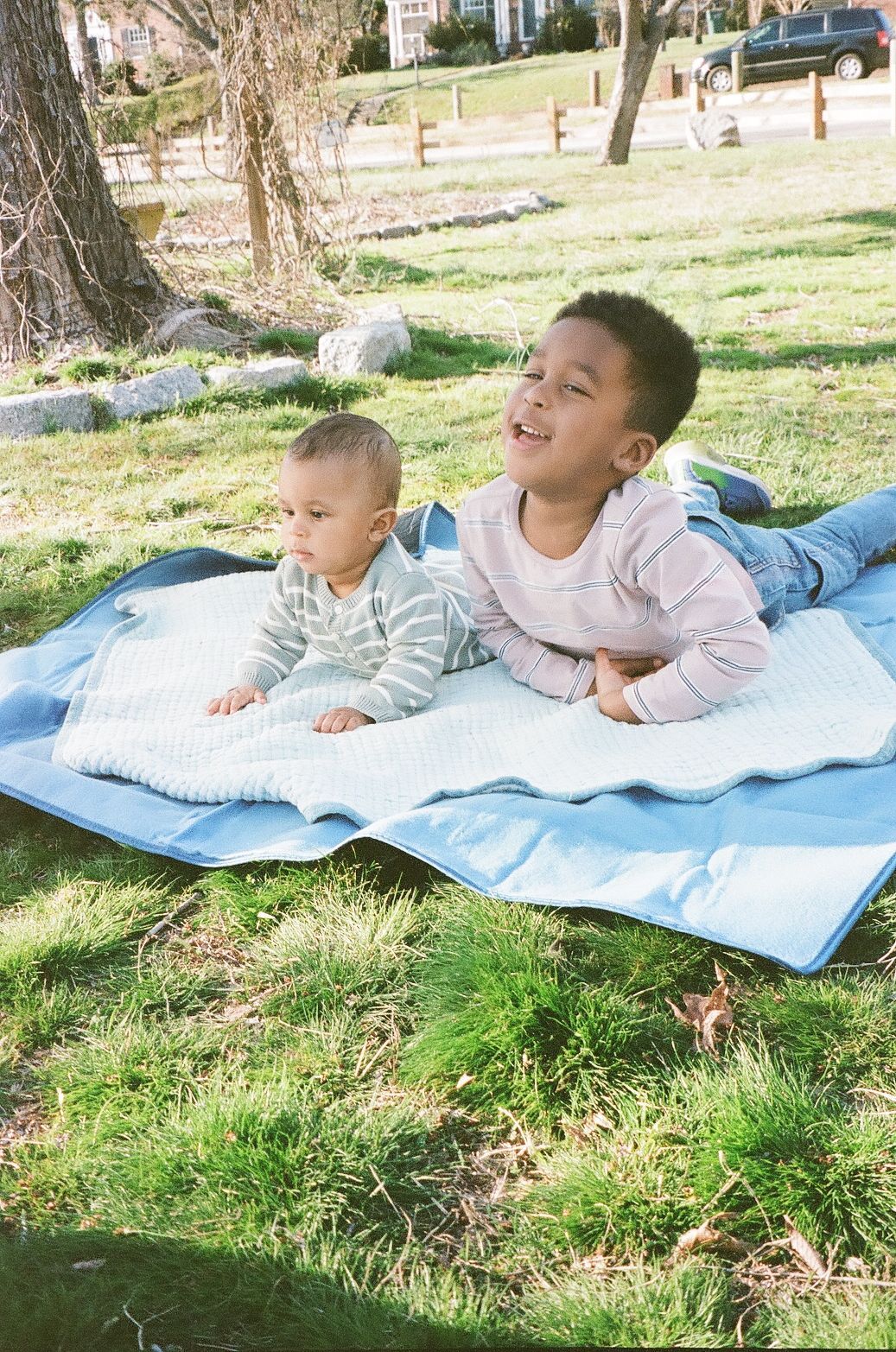 Two young children lying on a blue blanket on green grass outdoors on a sunny day.