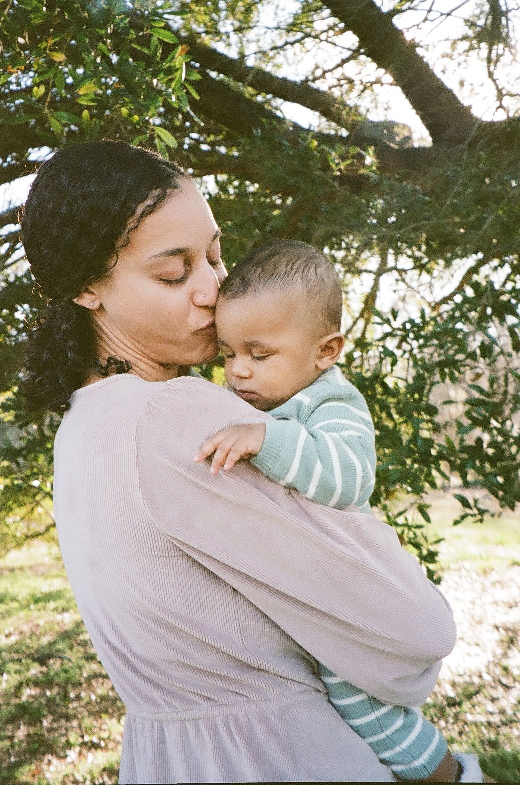 A mother tenderly kisses her baby outdoors, holding the infant close beneath a leafy tree in warm sunlight.