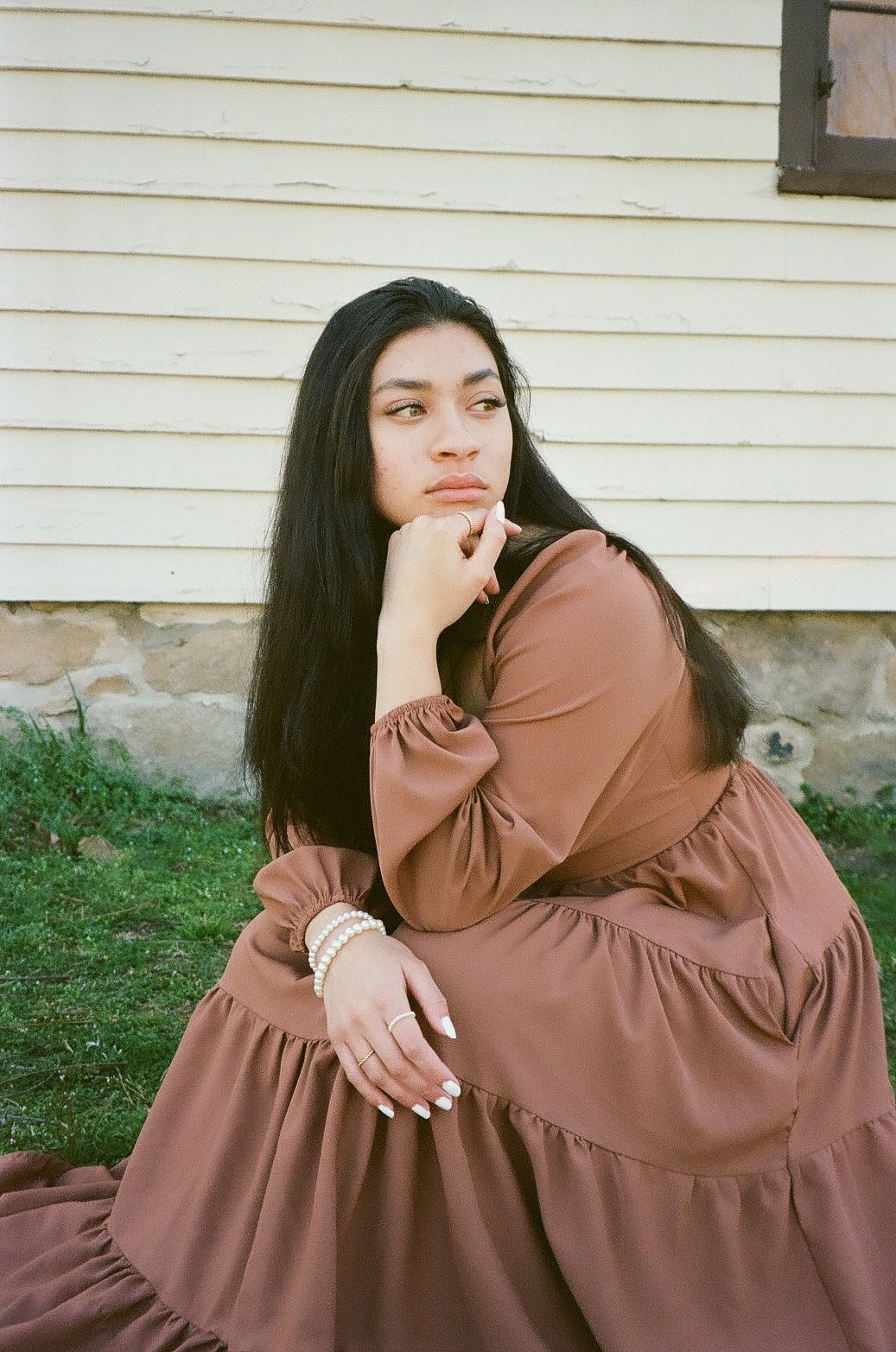Young woman in brown tiered dress sitting outdoors, resting chin on hand, wearing pearl bracelets.
