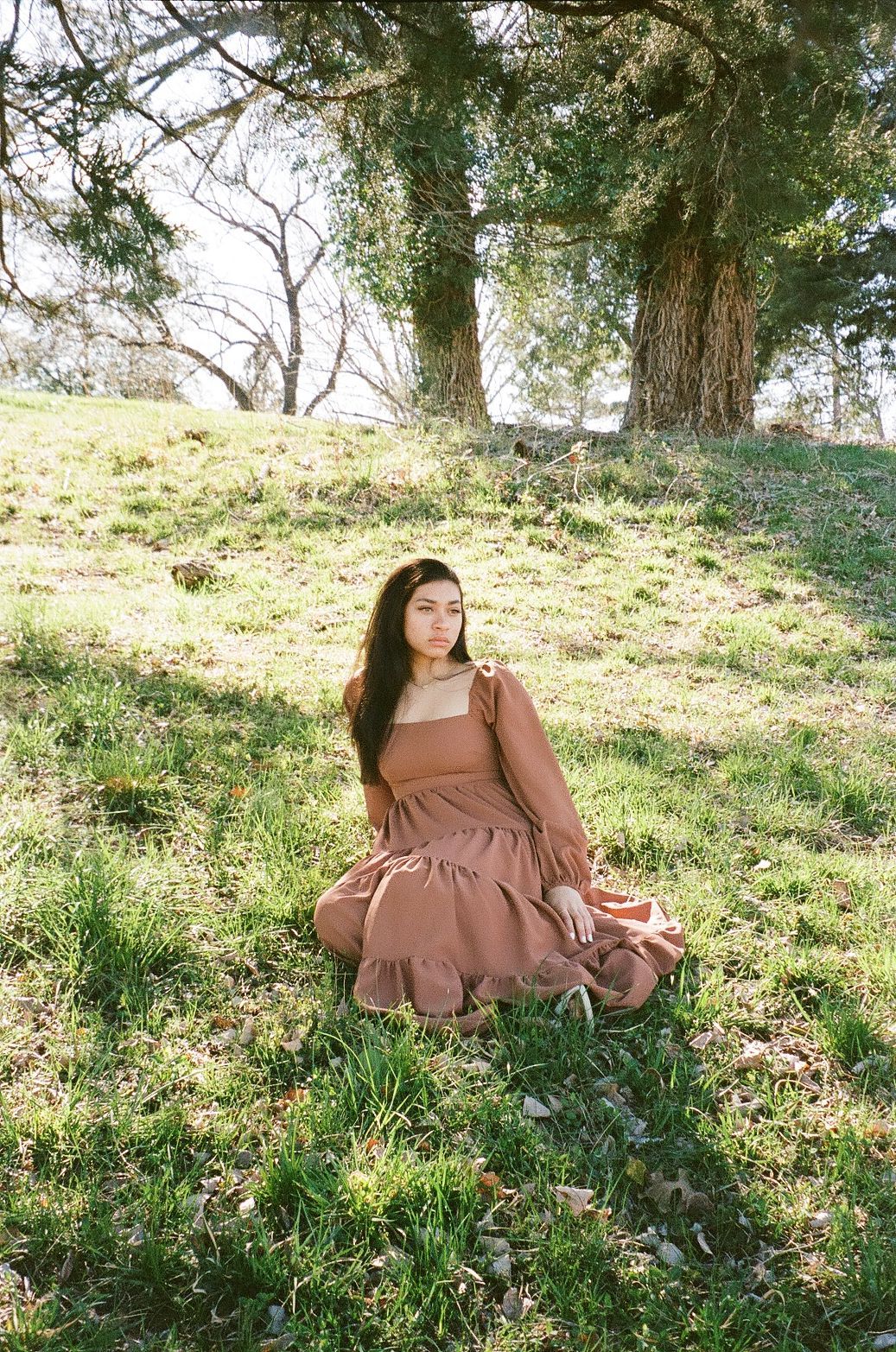 Young woman in a flowing rust-colored dress sitting on green grass beneath large trees in soft natural sunlight.