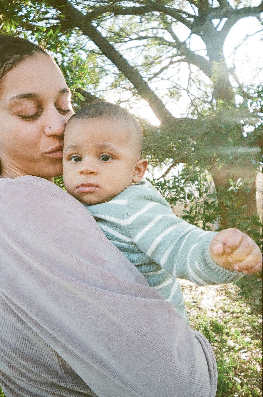 Parent and baby share a tender outdoor moment under dappled sunlight filtering through tree branches.
