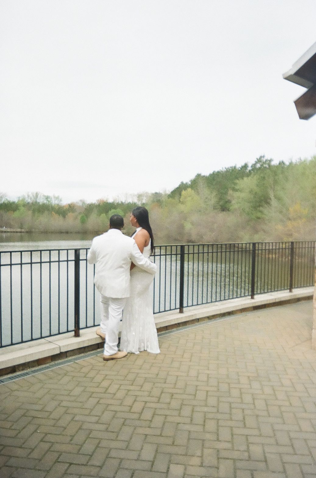 A couple in formal white attire stands at a lakeside railing on a brick patio, looking out over the water.