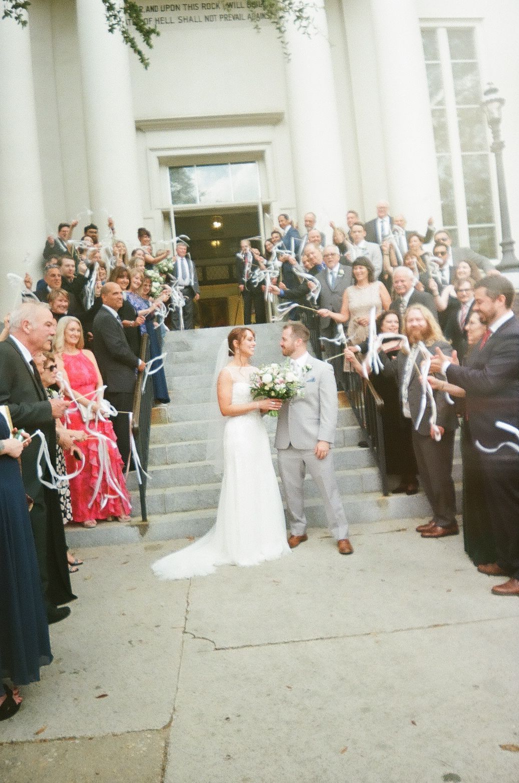Wedding couple stands on church steps surrounded by guests throwing white ribbons in celebration at outdoor ceremony.