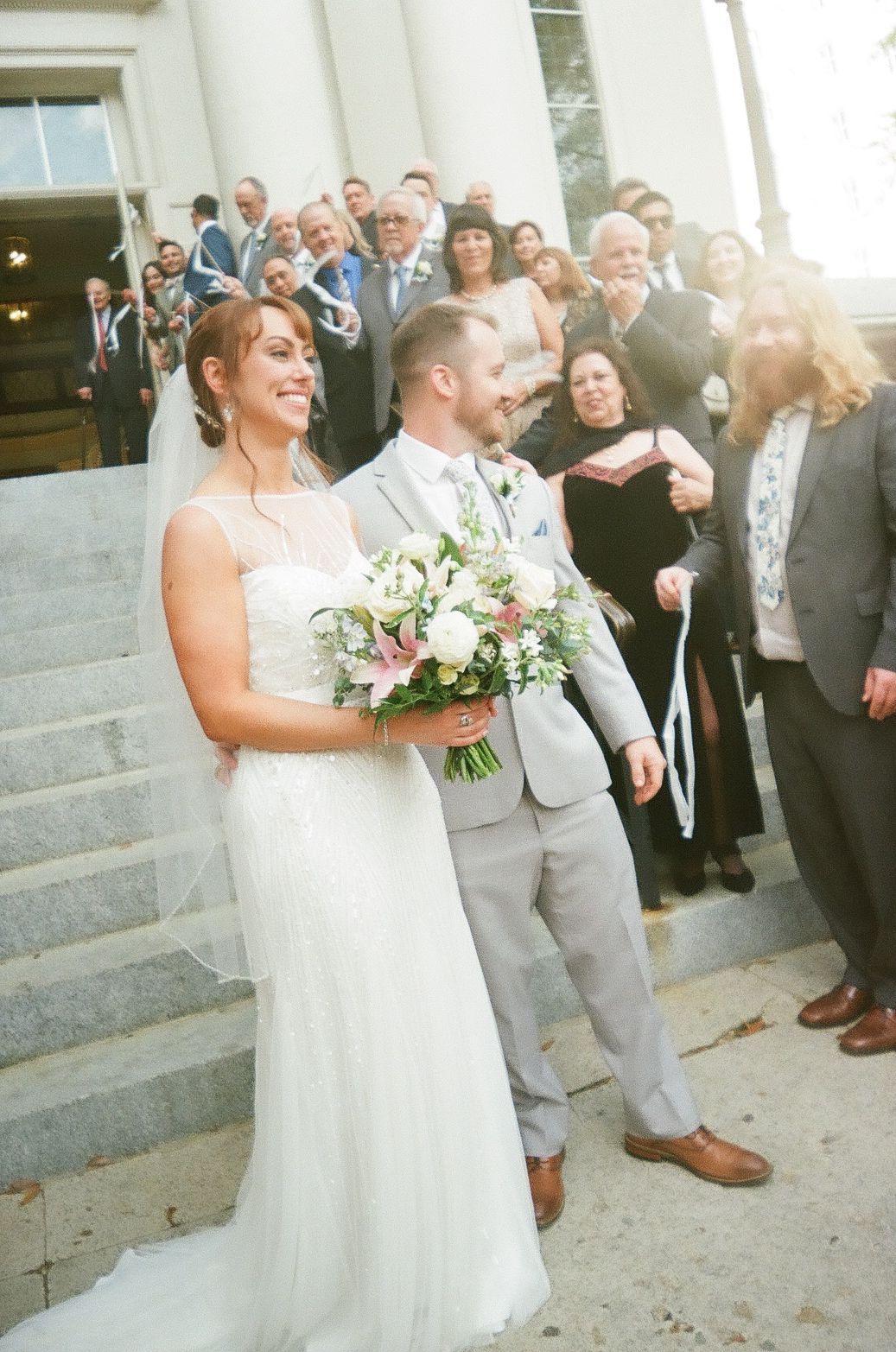 Wedding couple stands on steps with their wedding party gathered behind them in sunlight as bride holds white bouquet.