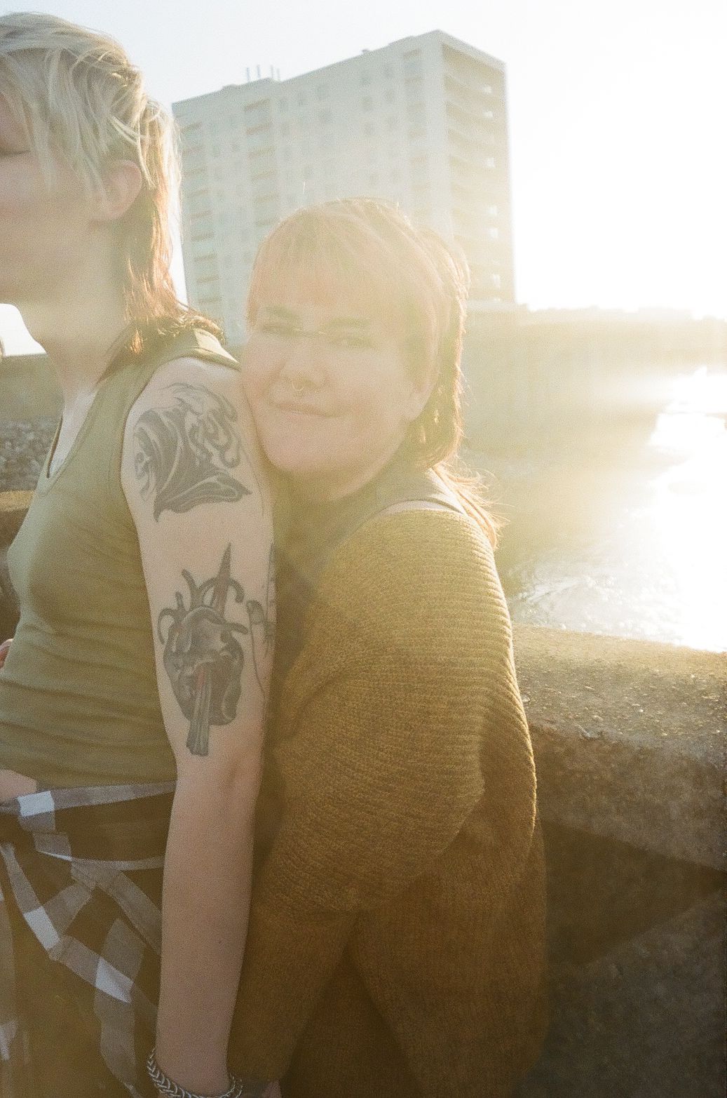 Two individuals sharing a sunlit moment outdoors, with one showing a visible arm tattoo and wearing a mustard sweater.
