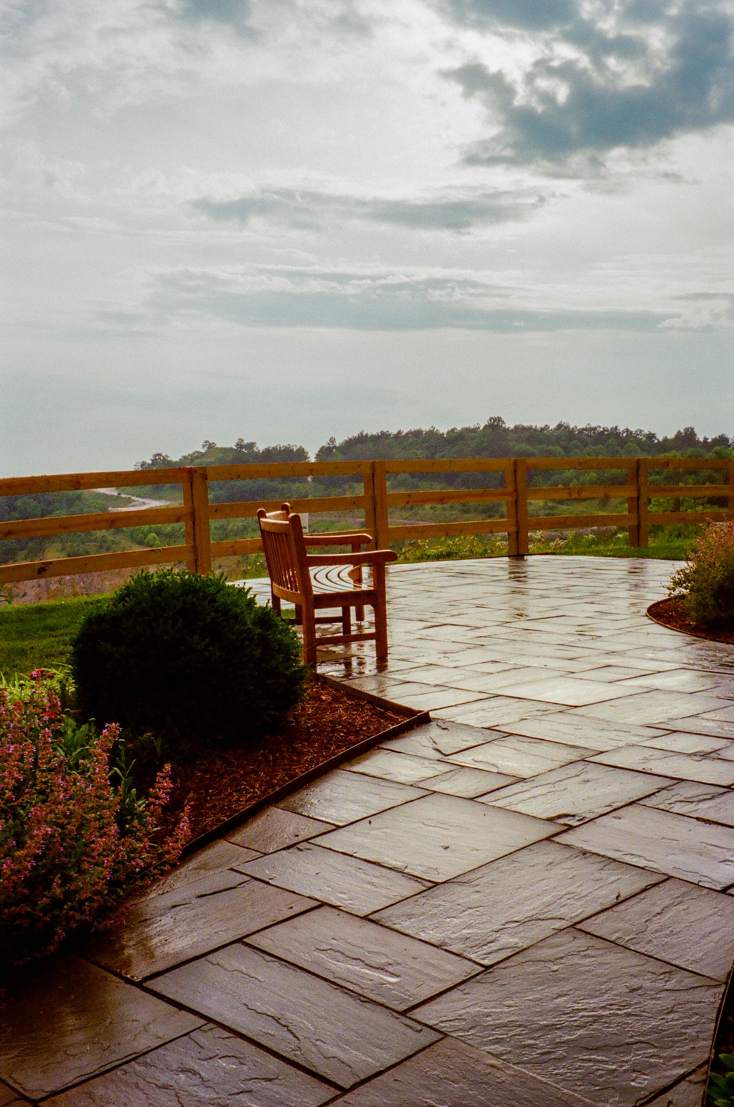 A stone patio with wooden railing overlooks a misty rural landscape with a lone wooden chair and landscaped garden beds.