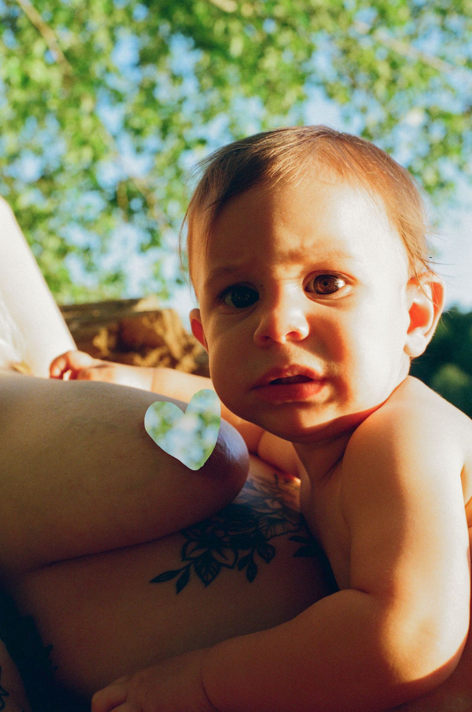 A baby with a curious expression being held by an adult with a floral tattoo outdoors under green trees.