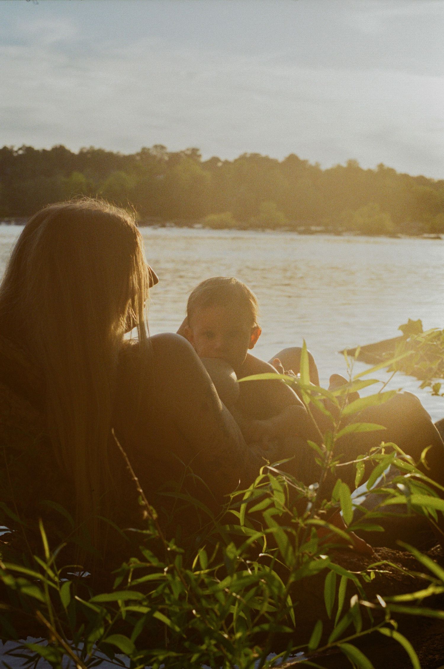 Two children sit together in golden sunset light by a tranquil river, surrounded by lush green plants.