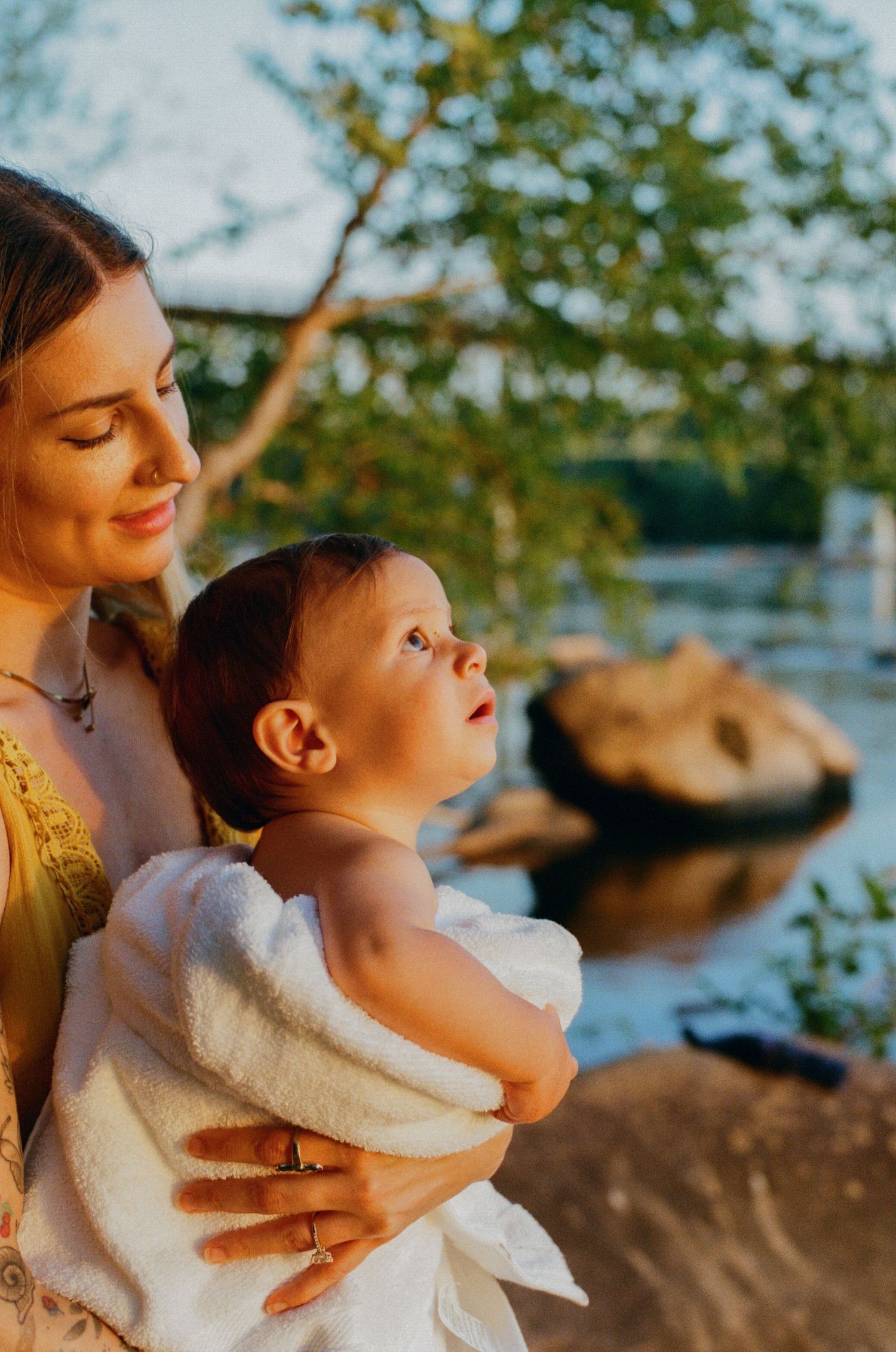 Mother holding baby wrapped in white towel by a sunlit lake at golden hour.