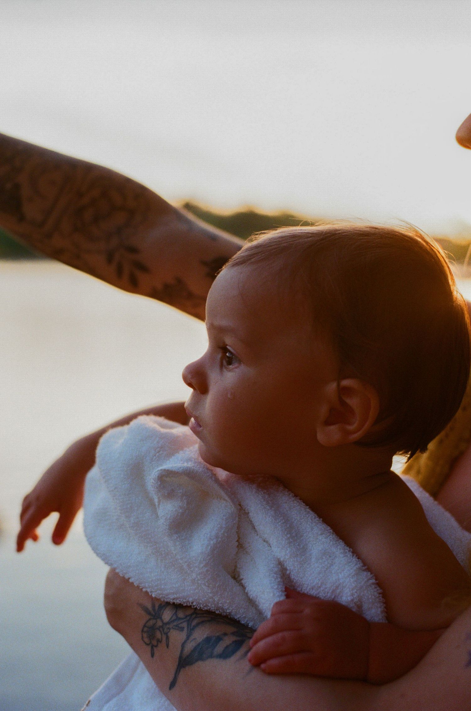 Tattooed parent cradling infant in white towel at sunset near water.
