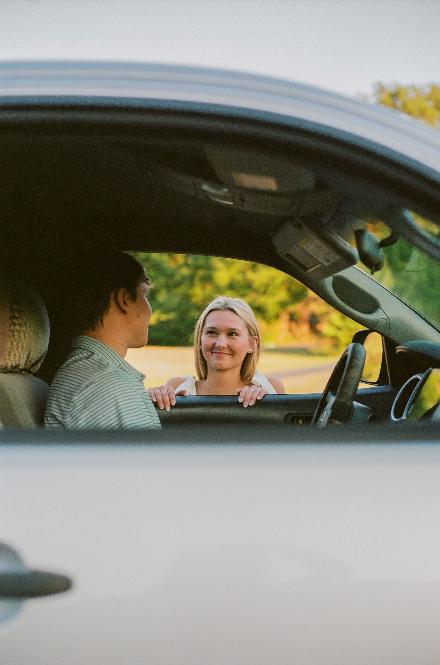 A smiling blonde woman leaning on a car door talking to a man in a striped shirt outdoors.