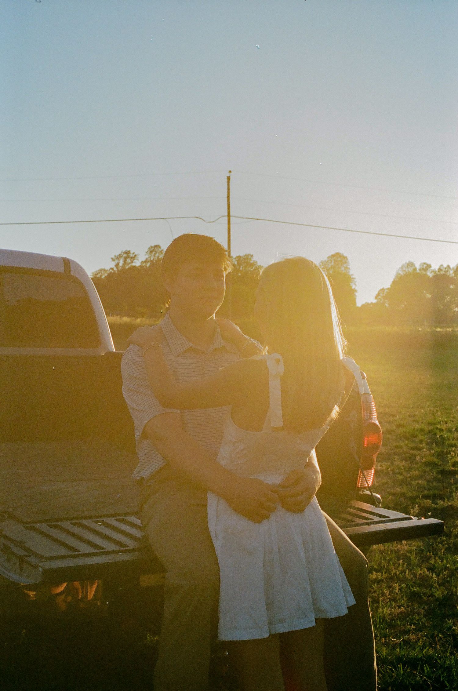 Couple embracing by a pickup truck at golden hour sunset in a rural field with warm lens flare.