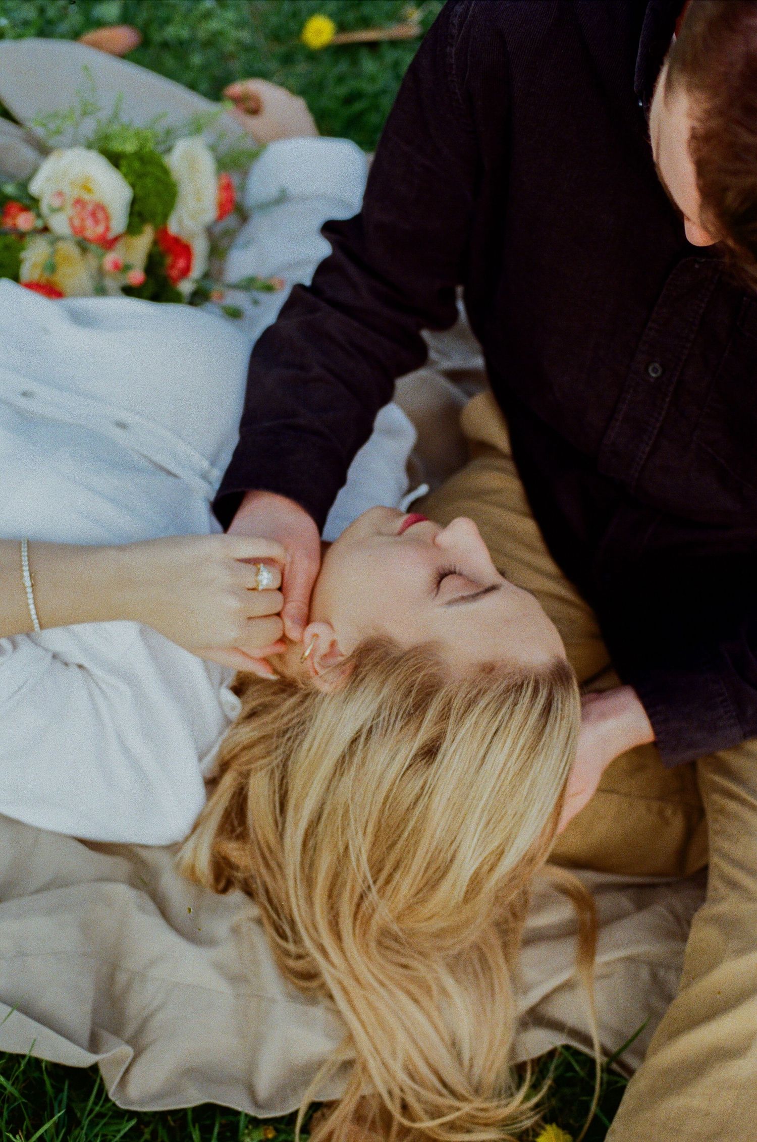 Couple sharing a tender moment on a blanket outdoors, woman lying down with engagement ring visible near colorful flowers.