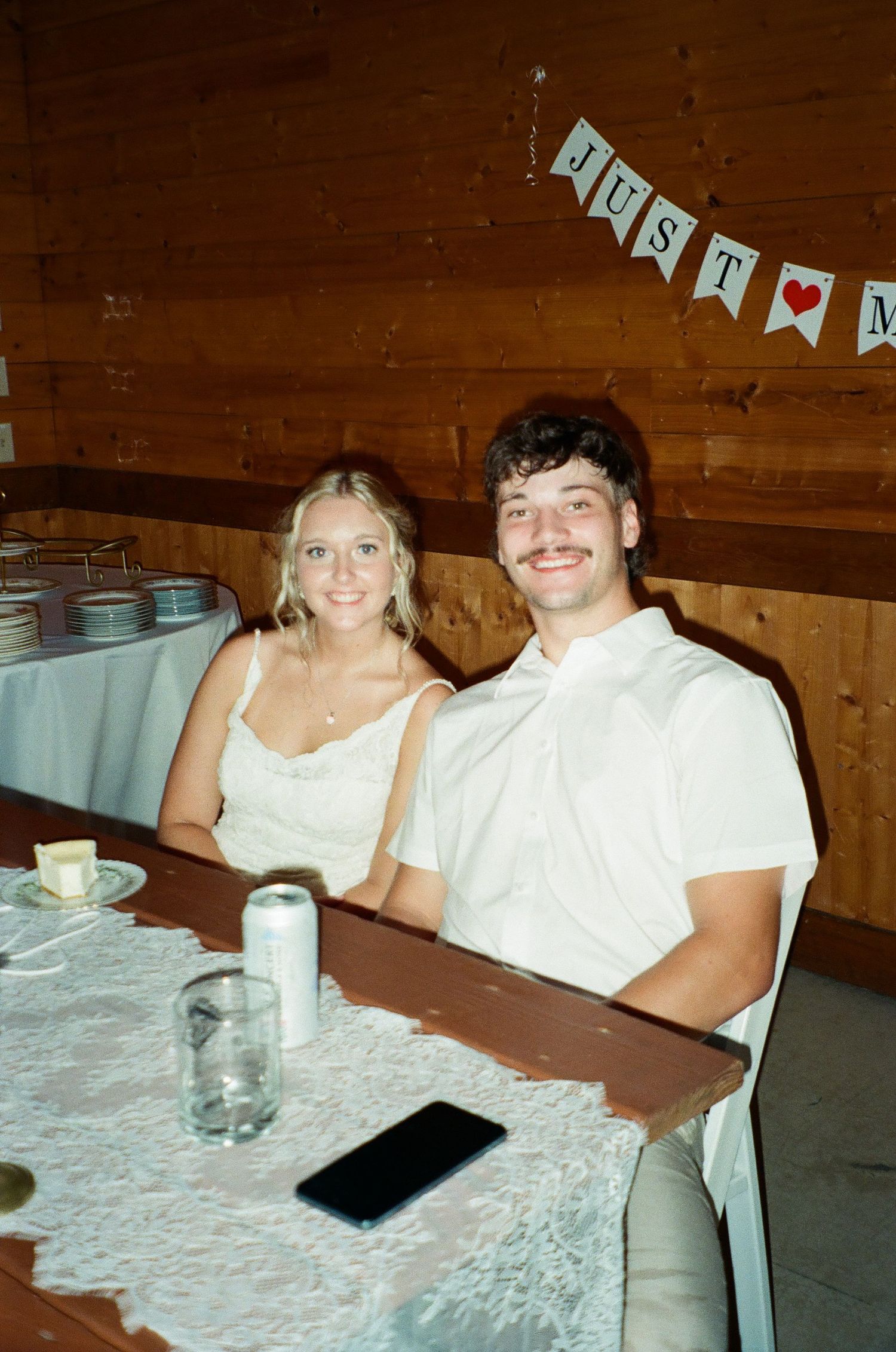 Couple smiling at a wedding reception table with a Just Married banner in a rustic wooden venue.