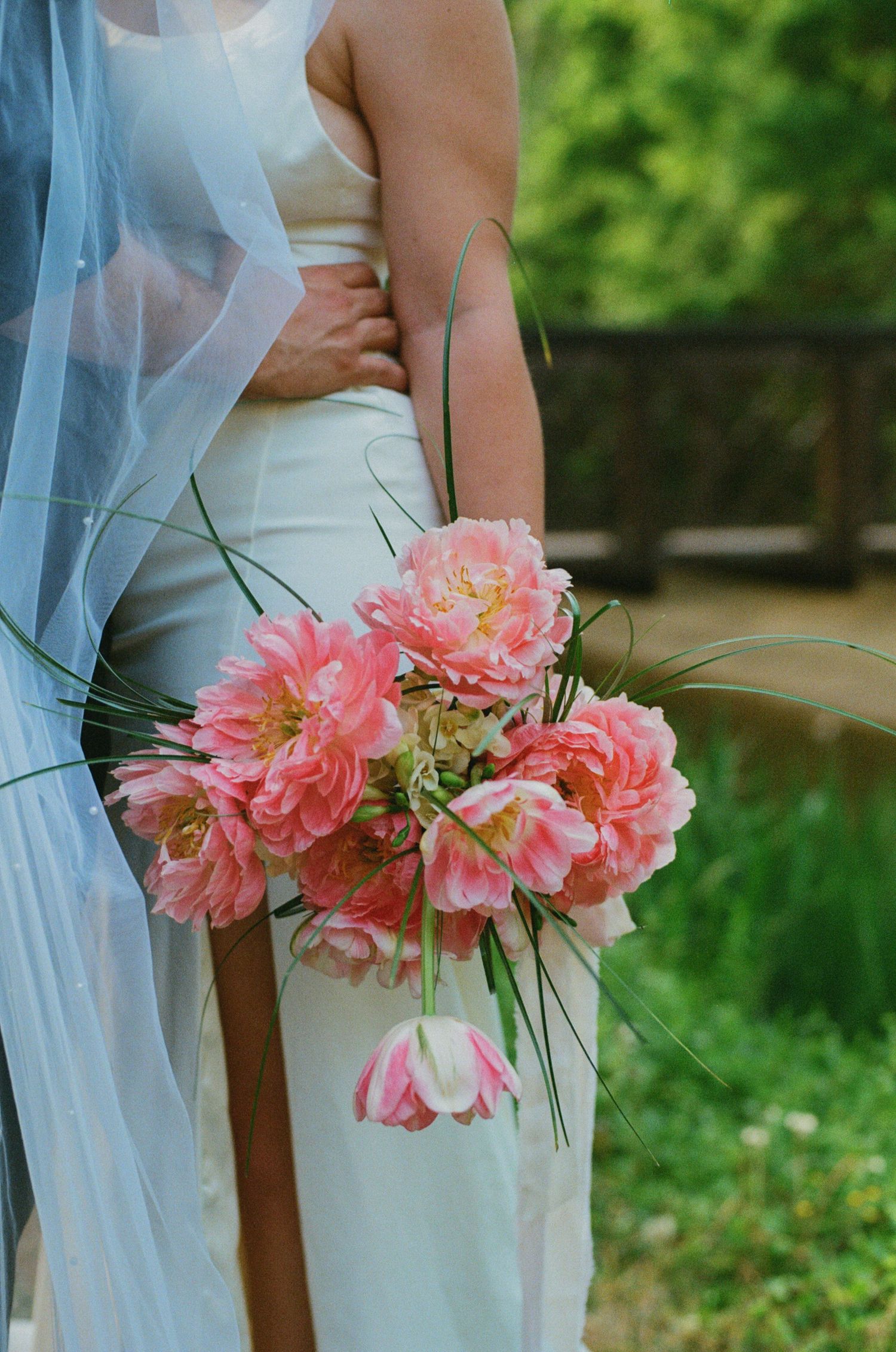 Bride in white gown holding a lush pink peony and tulip wedding bouquet with green grass accents outdoors.