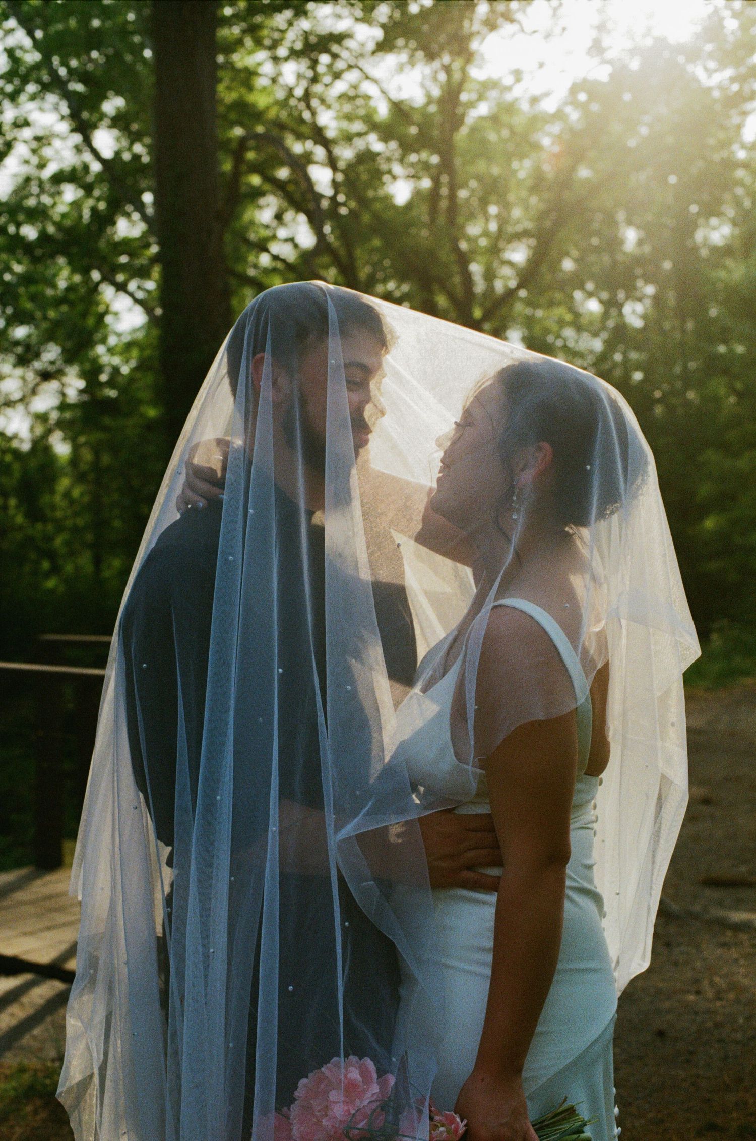 Bride and groom share a romantic kiss under a flowing veil in golden sunlight surrounded by lush green trees.