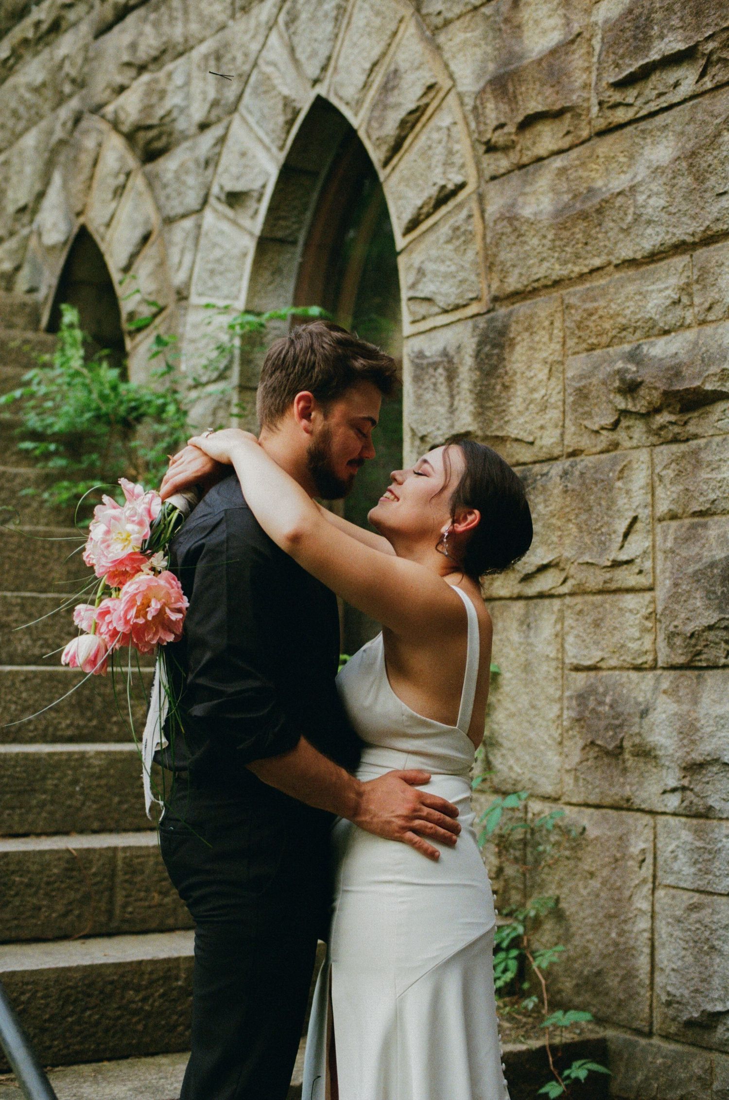Bride and groom embracing near stone arch wall, she holds pink peonies in a white gown, sharing a romantic moment.