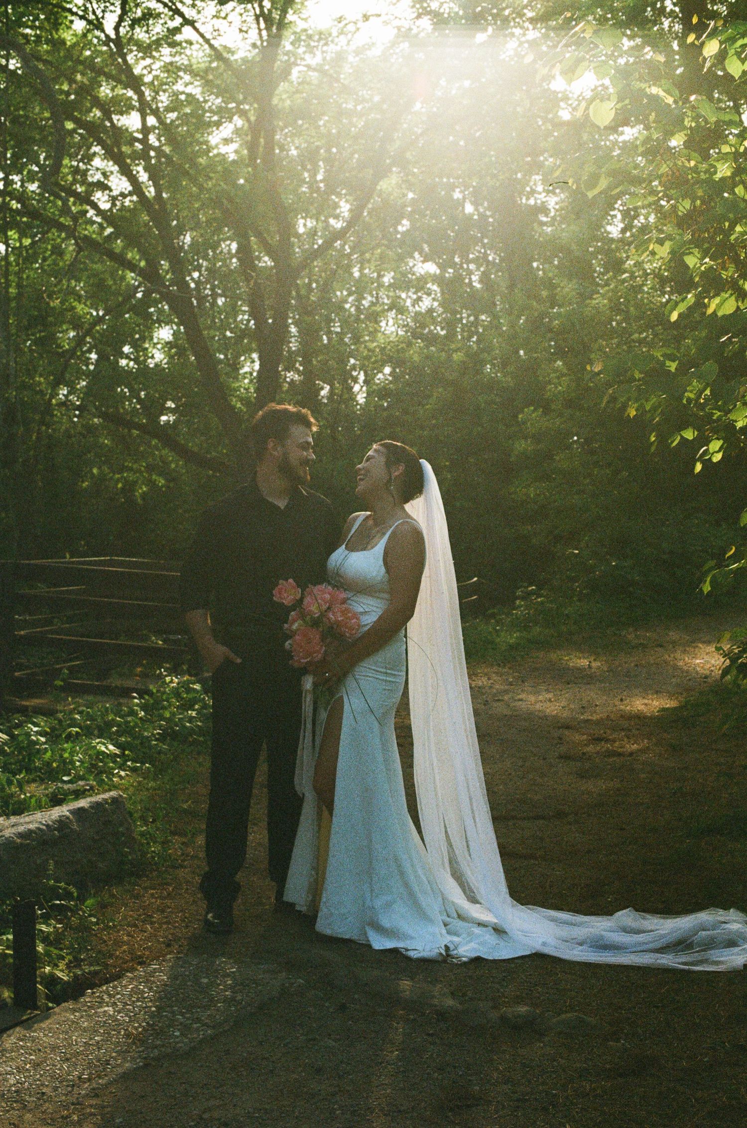 Bride in white gown and veil holds pink bouquet, gazing at groom in black, surrounded by lush green trees in golden sunlight.