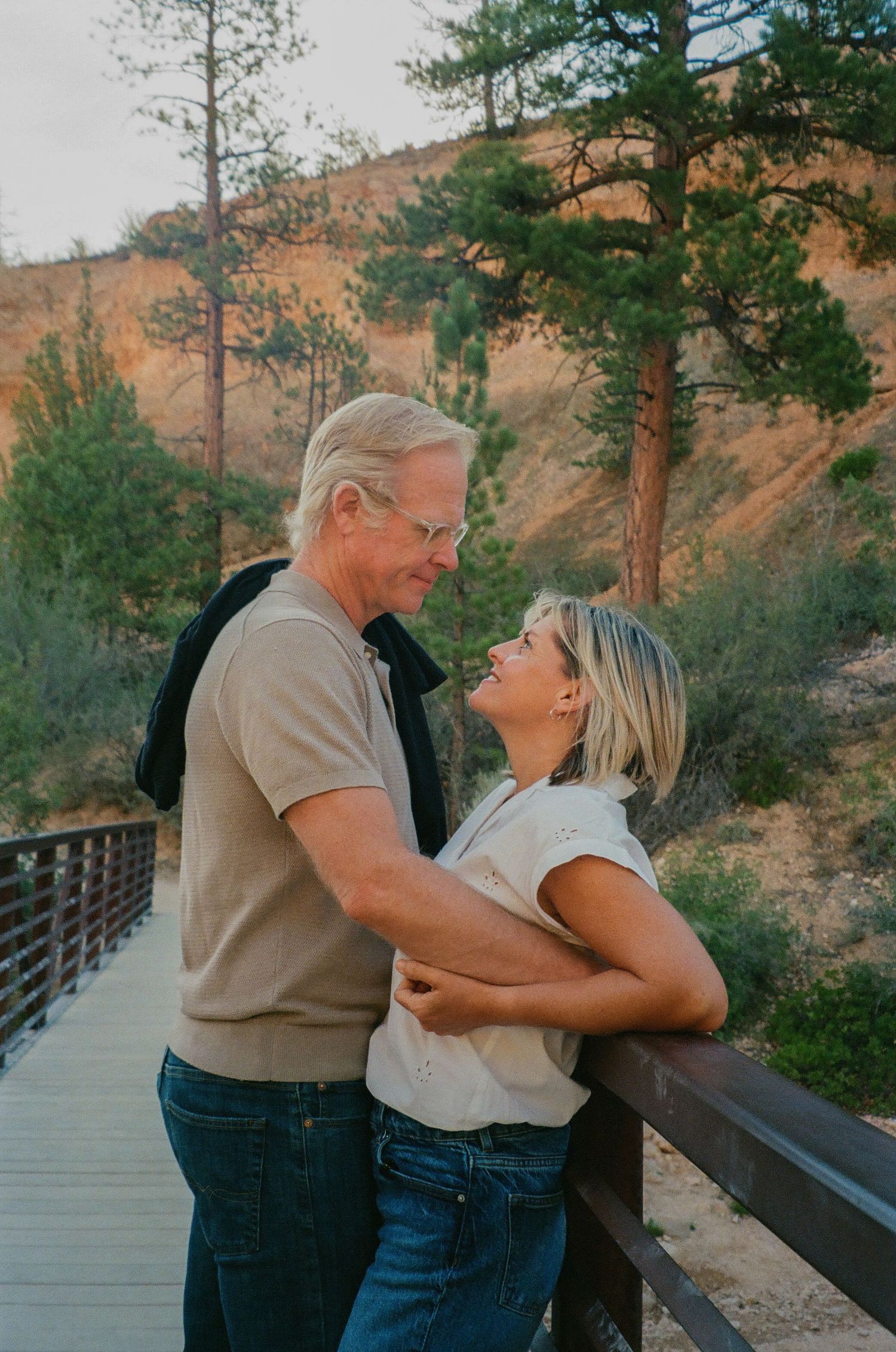 Couple embracing on a wooden bridge surrounded by pine trees and golden hillside landscape.