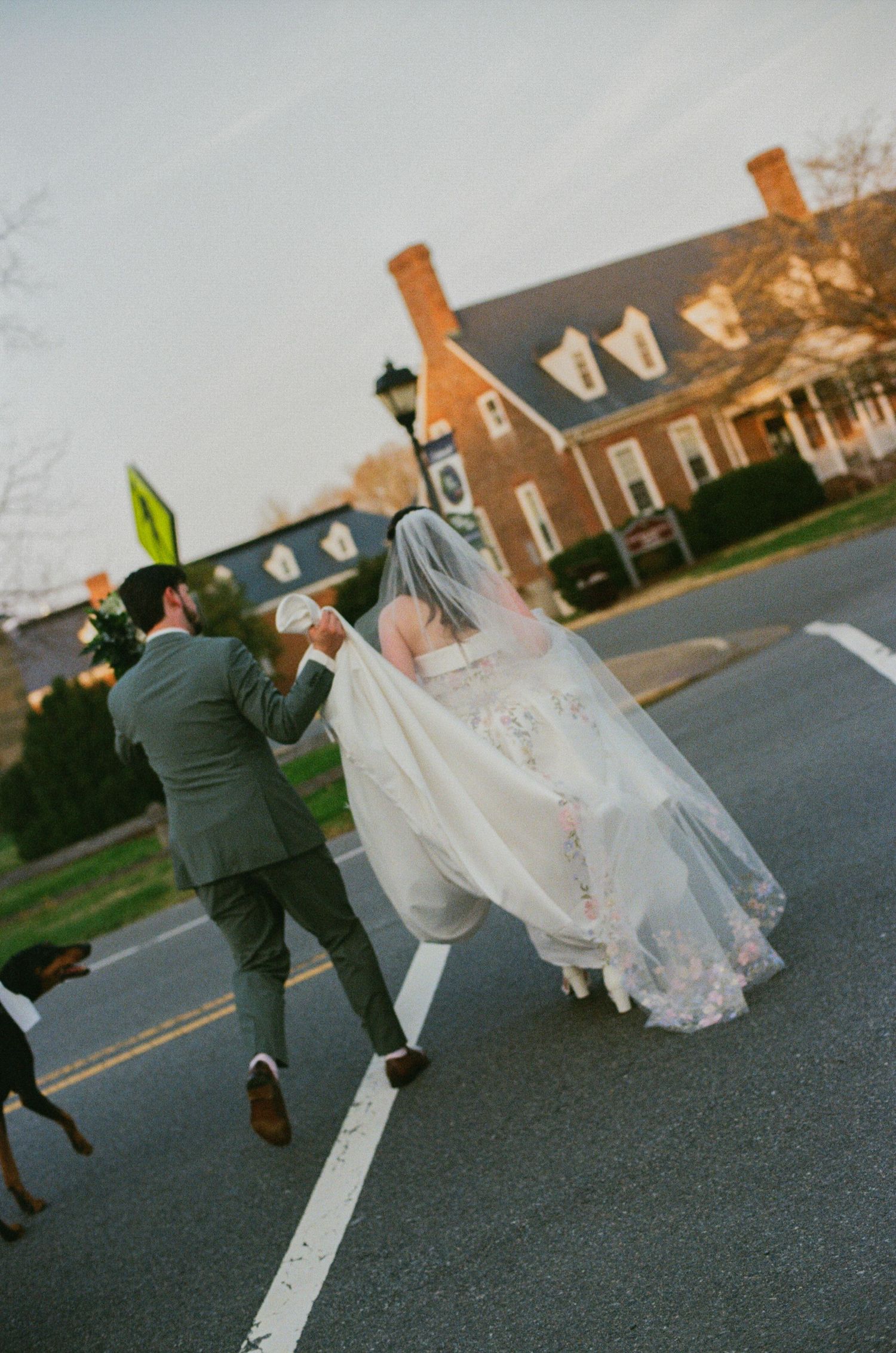 A bride and groom walk hand-in-hand across a street, her veil flowing, near a brick colonial building at dusk.