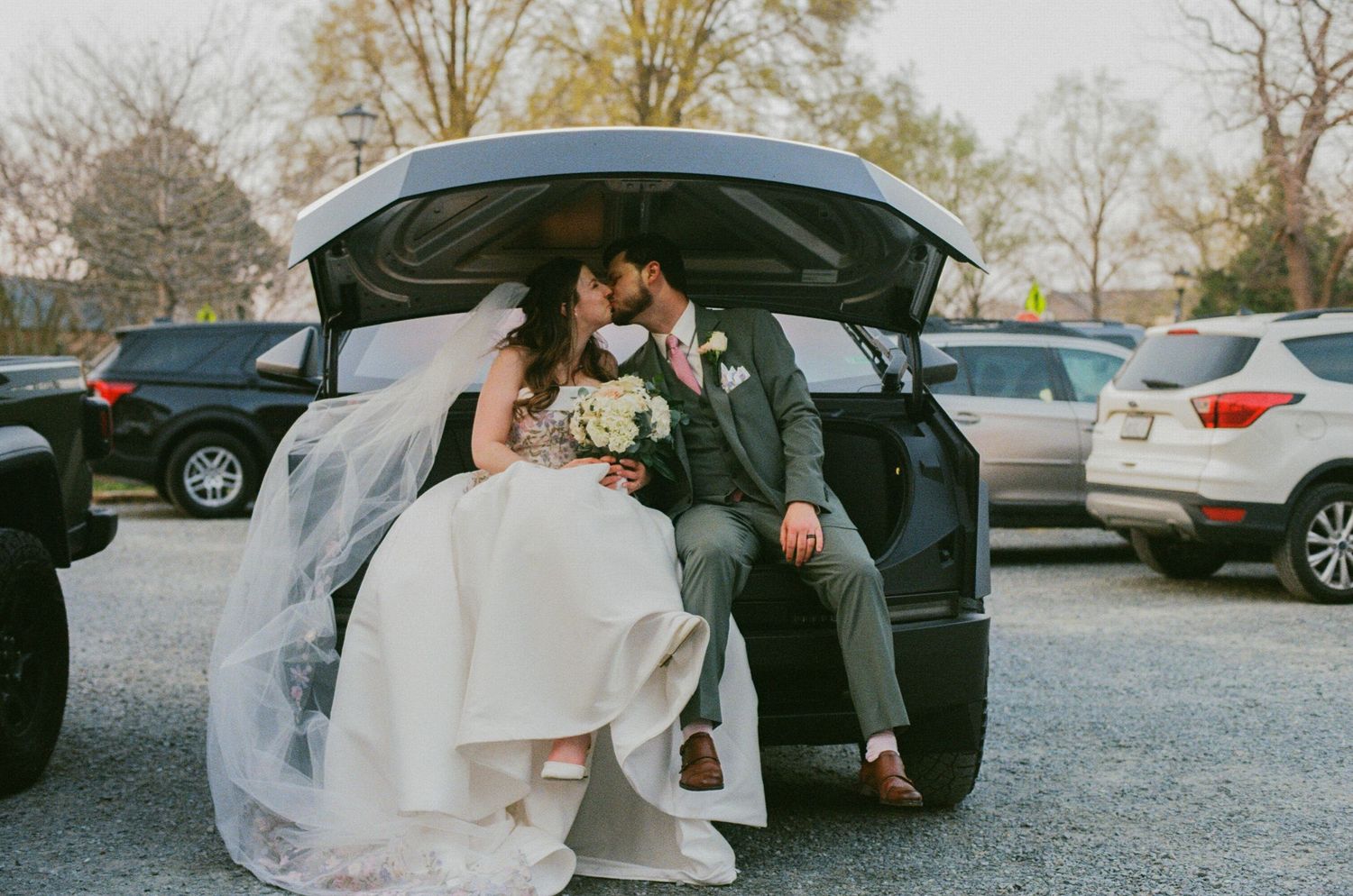 Bride and groom sitting in car trunk, sharing a kiss in parking lot, bride in white gown holding bouquet.