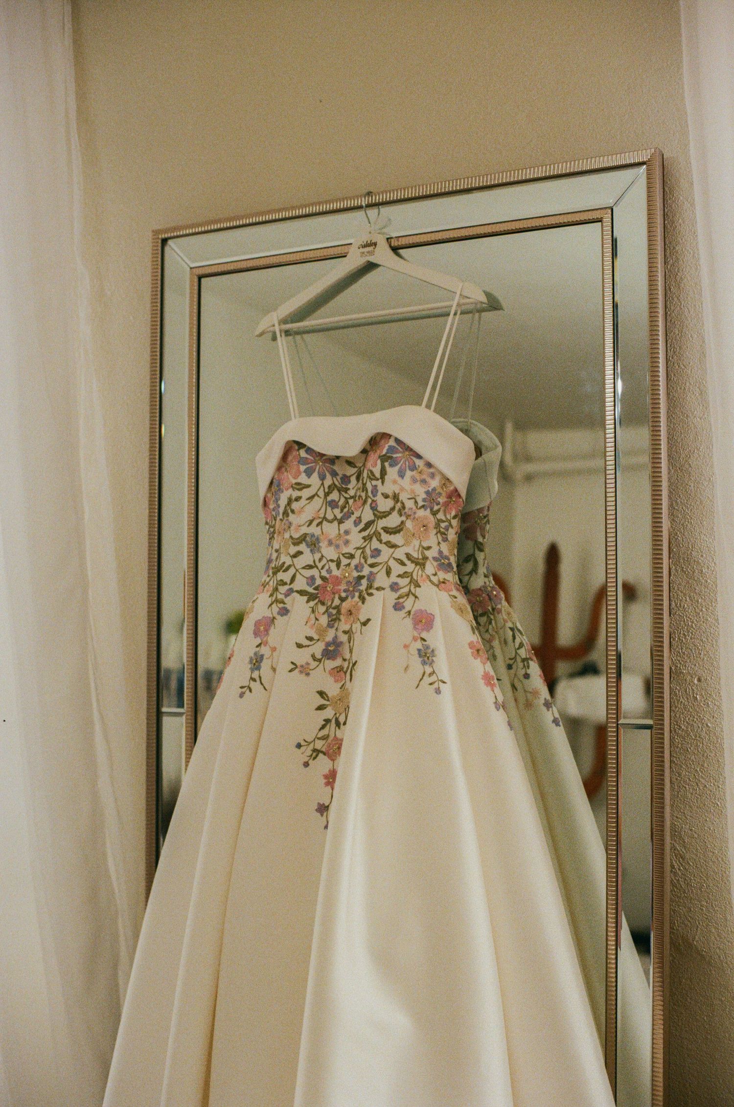 Elegant floral embroidered wedding gown with spaghetti straps hanging on a mirror in a bridal suite.