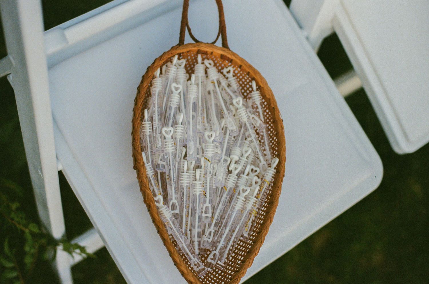 Vintage wooden tennis racket holding clear bubble wands on a white chair at an outdoor wedding ceremony.