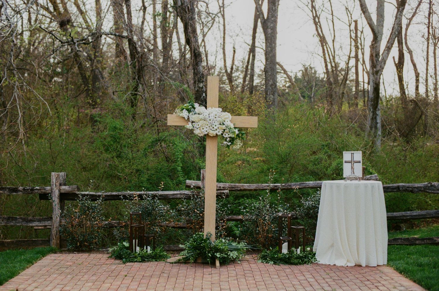 Rustic wooden cross adorned with white flowers at an outdoor wedding ceremony, surrounded by greenery and candles on a brick patio.
