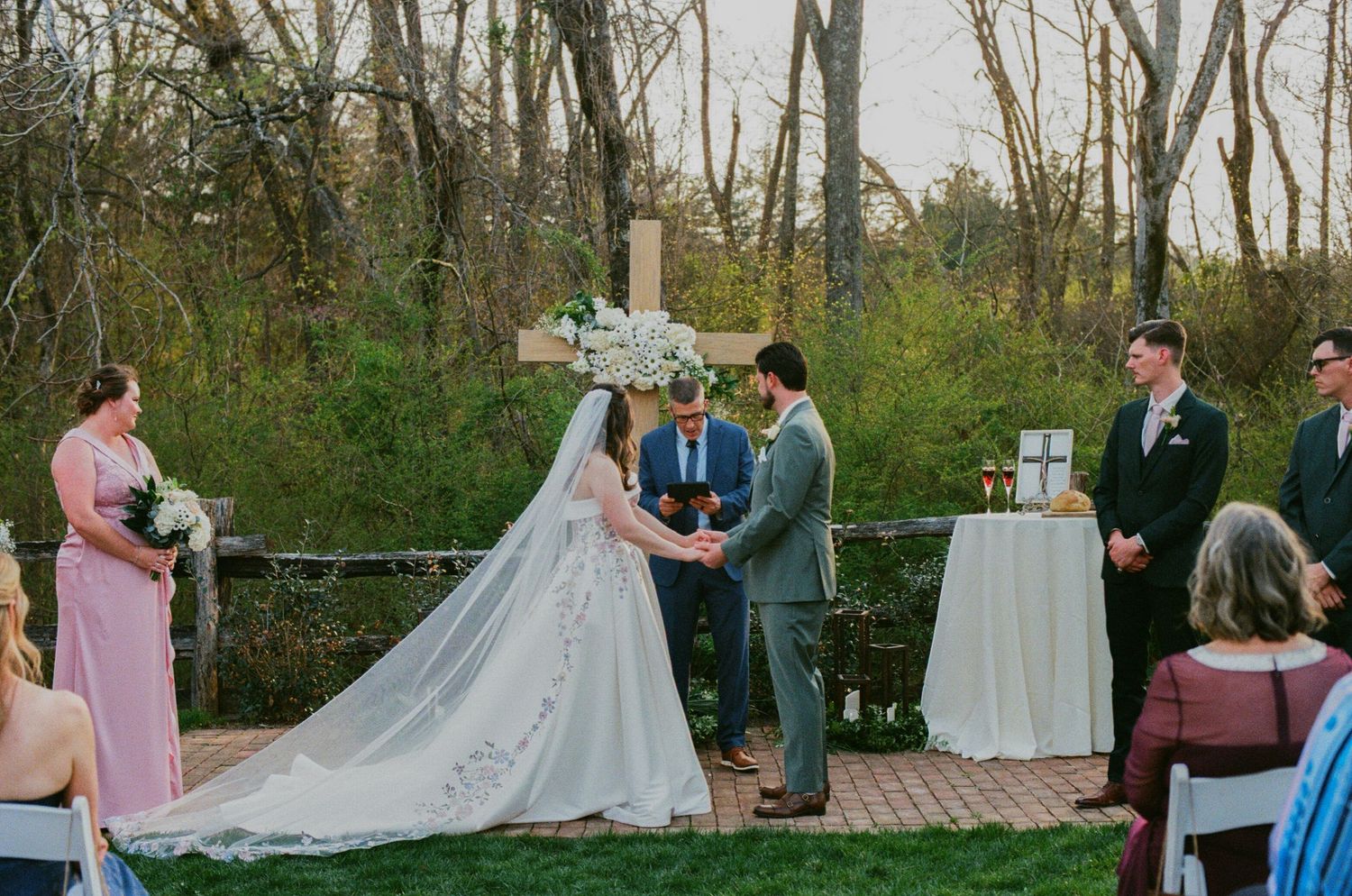 Outdoor wedding ceremony with bride in lace gown and veil, groom, and officiant before floral cross altar.
